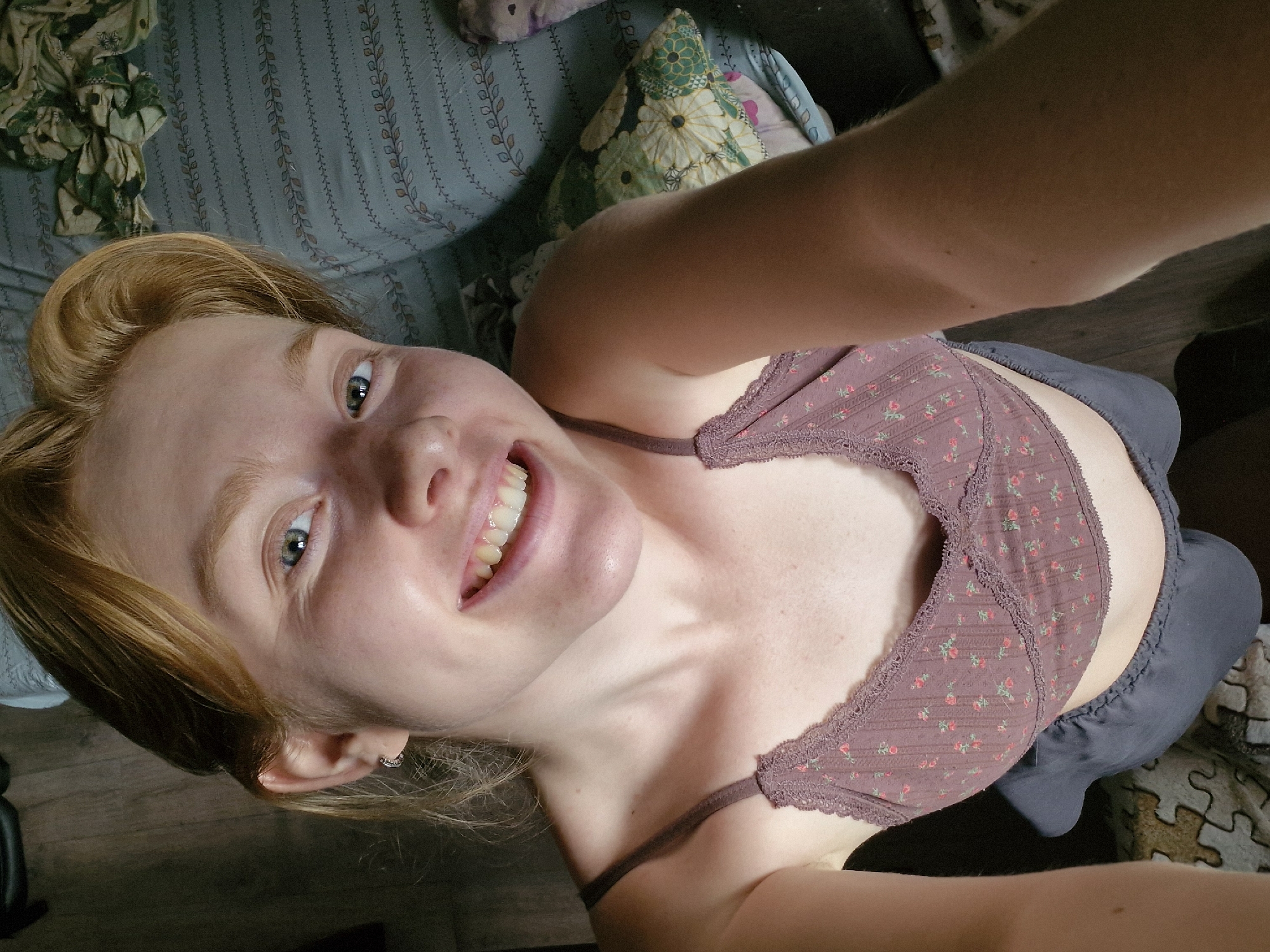 A light-skinned woman with short, auburn hair is taking a selfie. She is smiling and looking up at the camera, displaying her teeth. She is wearing a mauve bralette with a floral pattern and what appears to be a pair of grey shorts. The background includes a floral cushion, a blue leaf-patterned sofa, and a wooden floor.
This image does not contain any information to suggest it was taken in Yekaterinburg, Russia, or any specific location.