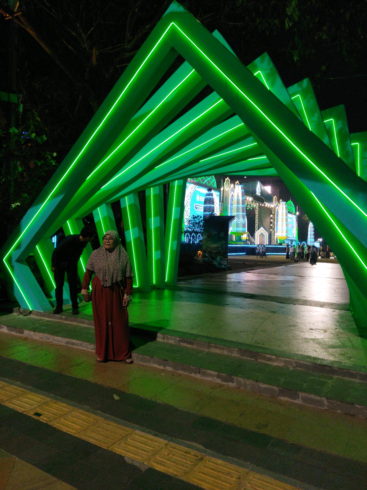 This is a nighttime, low-angle, medium shot photograph of an outdoor scene in Semarang, Indonesia, likely during a festive event, given the elaborate green neon light installation. In the foreground, a woman wearing a patterned hijab and a long, dark red dress stands on a paved walkway with tactile paving blocks. She is looking slightly to her right, with her hands clasped in front of her. Behind her, a man in a black shirt and pants is bending down, appearing to look at something on the ground or interact with the woman, though their interaction is not clear. The dominant feature of the image is a large, geometric structure made of green neon lights that forms an archway or tunnel. The neon lights are brightly lit, casting a green glow on the surroundings. Beyond the archway, a stage is set up with illuminated structures that resemble buildings with towers, possibly part of a religious or cultural celebration. Several other people are visible in the midground and background, walking and congregating near the stage. The ground surface is a mix of paved areas and steps, all illuminated by the green lights. The background trees are dark and silhouetted against the night sky. The overall atmosphere is festive and vibrant due to the prominent use of neon lighting.