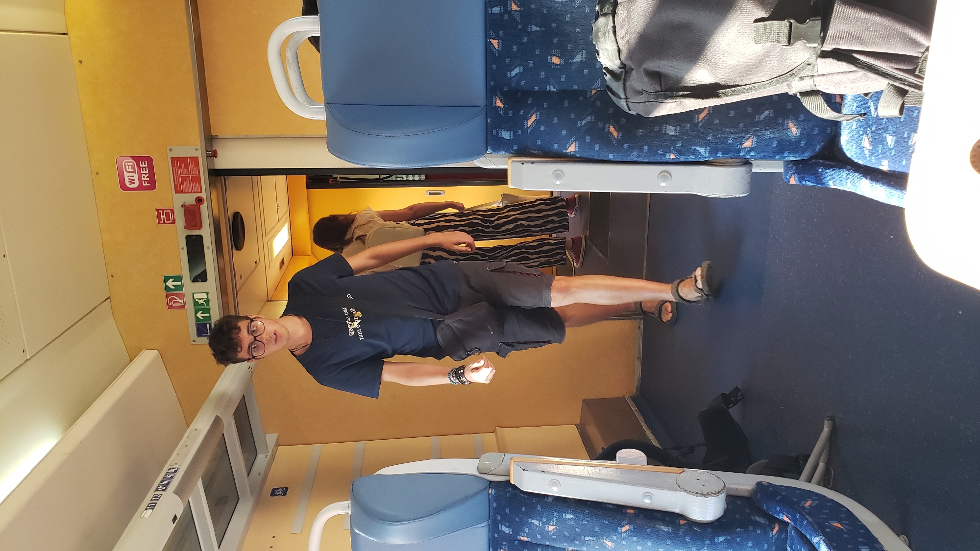 A young man with curly brown hair and glasses, wearing a dark blue t-shirt and dark shorts, walks down the aisle of a train car. He has a black crossbody bag slung over his left shoulder. He is barefoot. Behind him, a woman with long brown hair, wearing a light-colored top and black and white striped pants, walks away from the camera. The train car has blue seats with a patterned design and a yellow wall. There are signs on the wall, including a "Wi-Fi FREE" sign. The floor of the train car is dark blue.