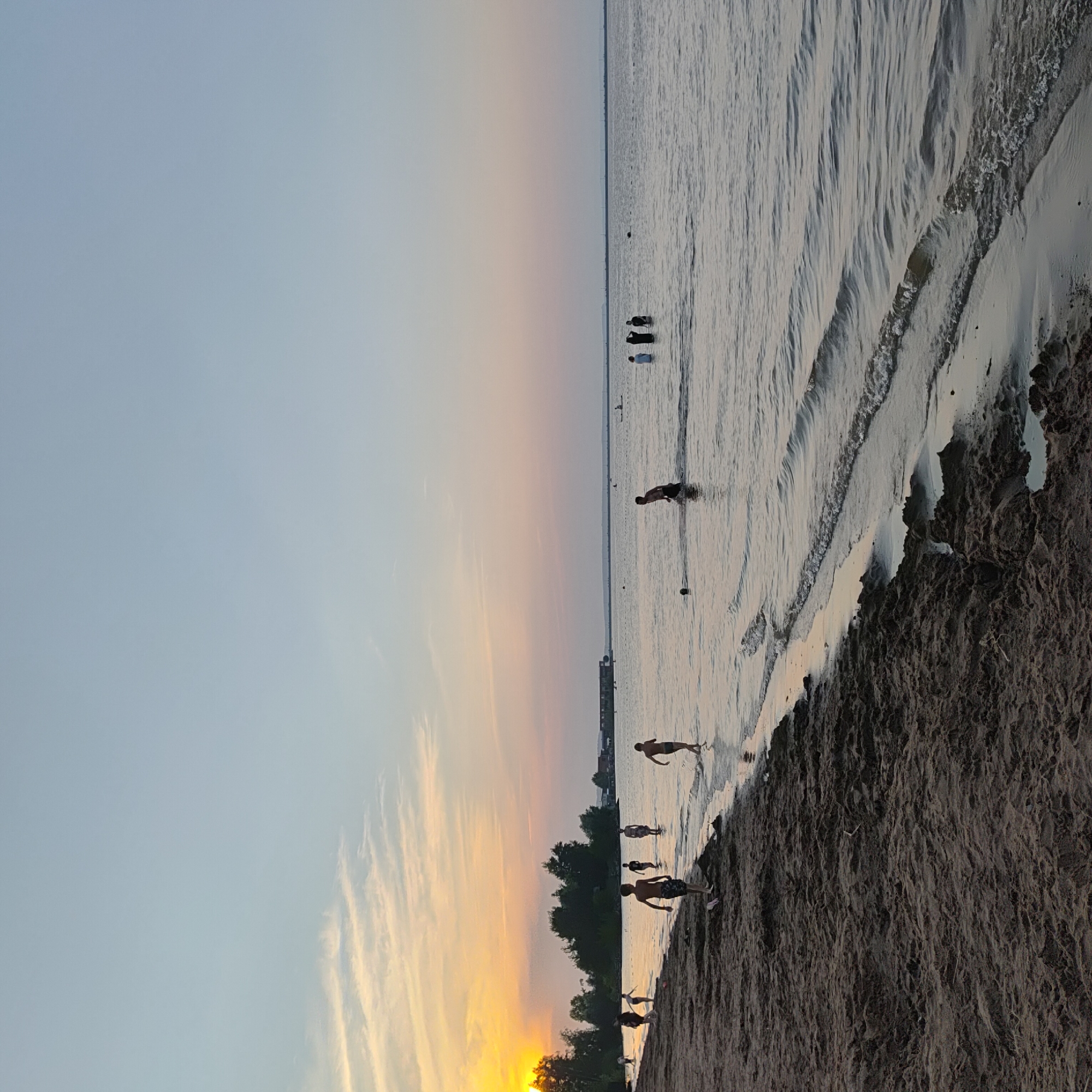 The image captures a sunset scene at a beach, likely in Chaikovsky, Russia, given the context. The sky is a gradient of soft blues and pinks, with wispy clouds illuminated by the setting sun on the left. The sun itself is a bright, warm orange orb peeking through a line of trees in the distance.
In the foreground, a sandy beach is visible, with footprints and ripples in the wet sand near the water's edge. Several people are scattered across the scene, both on the beach and in the shallow water. On the beach to the left, a group of figures are walking. Further into the water, several individuals, appearing to be children or teenagers, are wading and playing. One figure in the middle ground is walking towards the viewer, and another is further out in the water. In the distance, there are more figures in the water, some appearing to be on what might be a paddleboard or small boat. A low-lying building structure is visible across the water in the far distance. The water itself is calm, with gentle waves lapping the shore. The overall atmosphere is serene and summery.