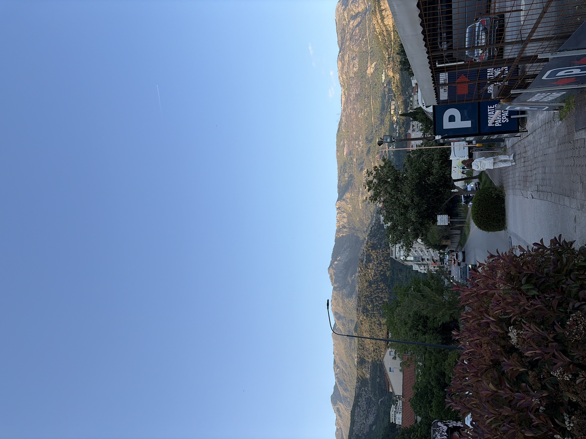 This bright, daytime photograph captures a street scene in Budva, Montenegro, with imposing mountains as a backdrop. The sky is a clear, vibrant blue, traversed by a faint airplane contrail.
In the foreground, a paved sidewalk and street are visible, with several vehicles, including parked cars and a white bus, along with a streetlight. A person dressed in white clothing stands on the sidewalk near a prominent blue sign featuring a large white "P" and the text "PRIVATE PARKING SPACE". Other partially obscured signs display arrows and the words "PRVA TAXI".
To the right, green and reddish-purple bushes with small white flowers line the path. Further along the street, multi-story buildings, some with red-tiled roofs, are interspersed with greenery, including mature trees.
The background is dominated by rugged, rocky mountains densely covered in vegetation, showing hints of human development like buildings and power lines on their slopes. The sunlight is strong and direct, suggesting mid-day or early afternoon. The scene depicts a sunny, clear day in an urban area nestled against a dramatic natural landscape.