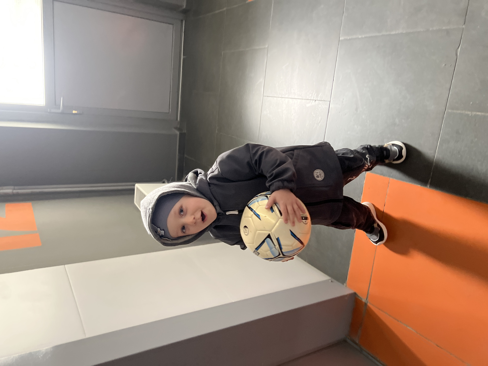 A young child, estimated to be around 3-5 years old, is holding a soccer ball in what appears to be an indoor corridor. The child is wearing a dark grey hooded jacket and dark pants. The flooring is a combination of grey tiles and orange tiles. A white wall with a grey baseboard is visible on one side. A grey door or window frame is seen in the background. An orange Nike logo is visible on the wall in the back ground.
Given that the image was captured in Mytishchi, Russia, based on other data, it is possible the scene depicts an indoor athletic facility or sports complex within the city.