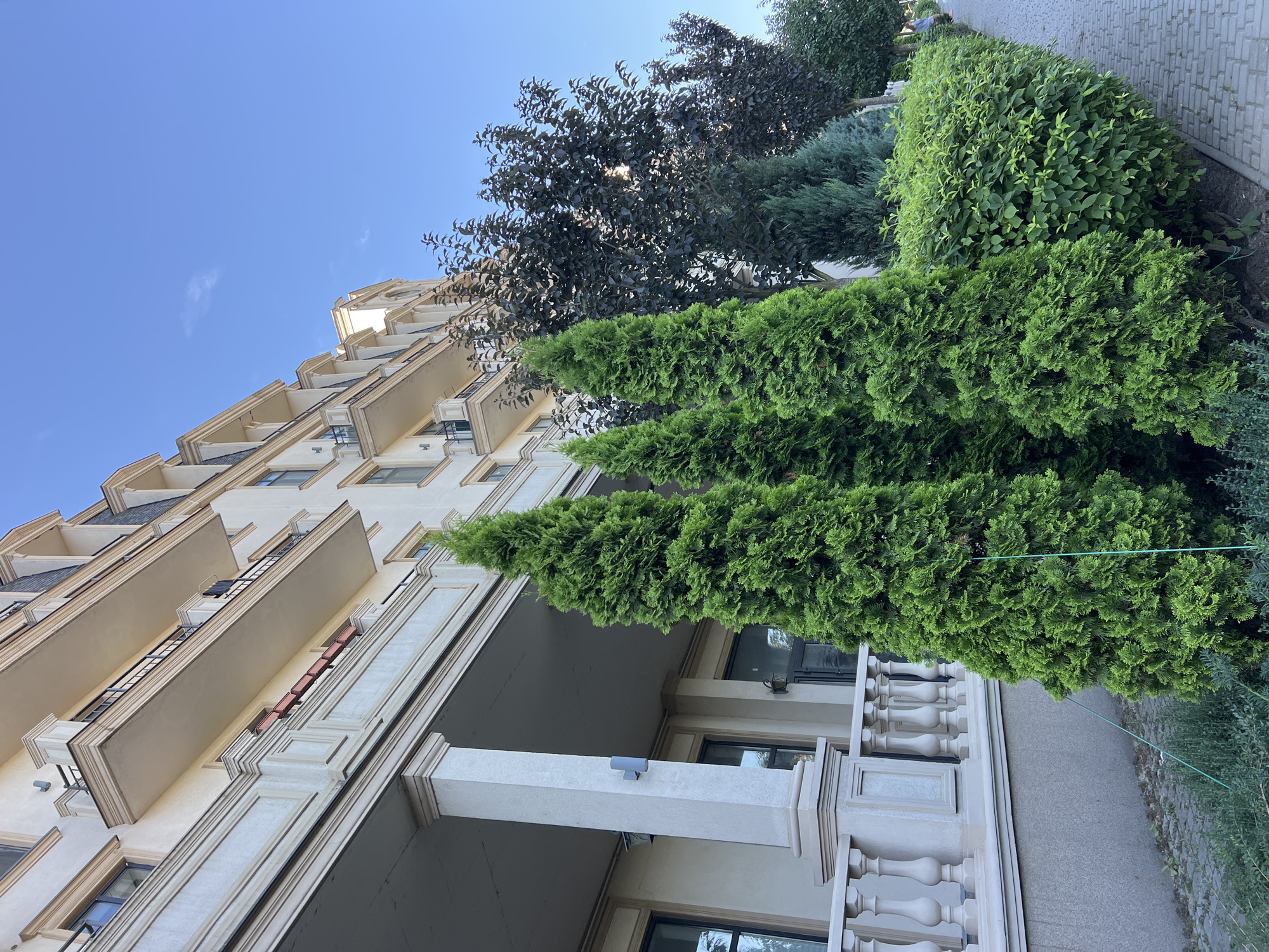In Fontanka, Ukraine, a multi-story building with balconies and dormer windows stands under a clear blue sky. In front of the building, a paved walkway runs alongside manicured greenery, including several tall, vibrant green cypress trees and a neatly trimmed hedge. The building's facade is light-colored with decorative architectural elements.