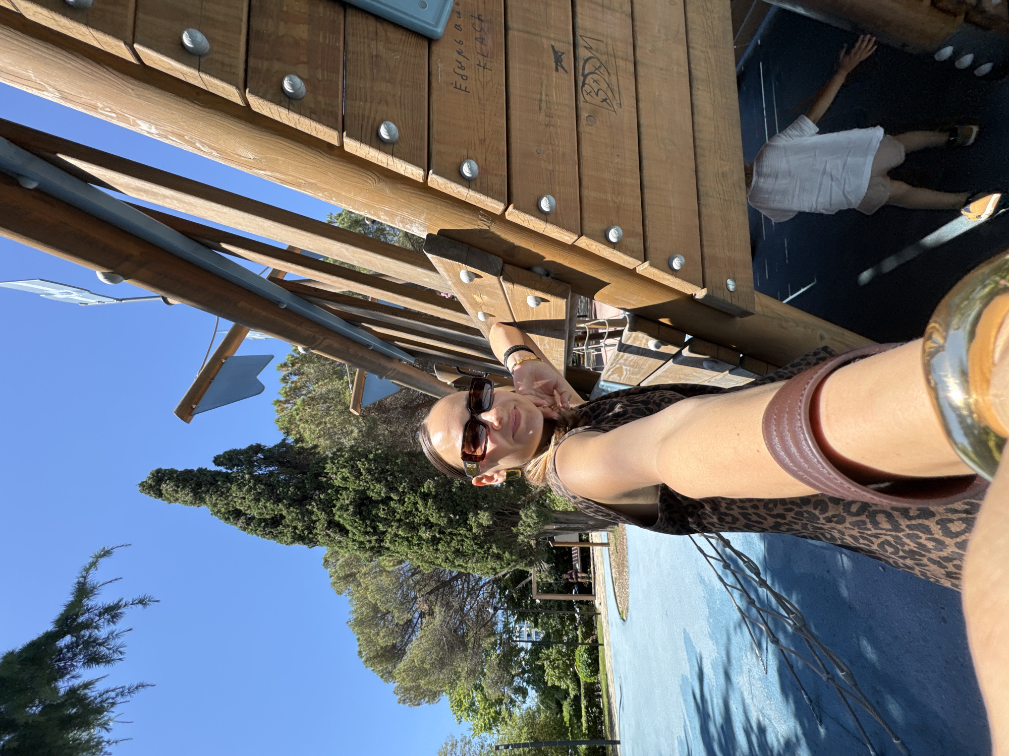 A woman with sunglasses and a leopard print dress is taking a selfie outdoors in a park. She is leaning against a wooden structure, possibly a playground. In the background, there are trees, a blue sky, and a child wearing a white t-shirt and shorts. There is graffiti on the wooden structure. The location is the city of Sochi, Russia.