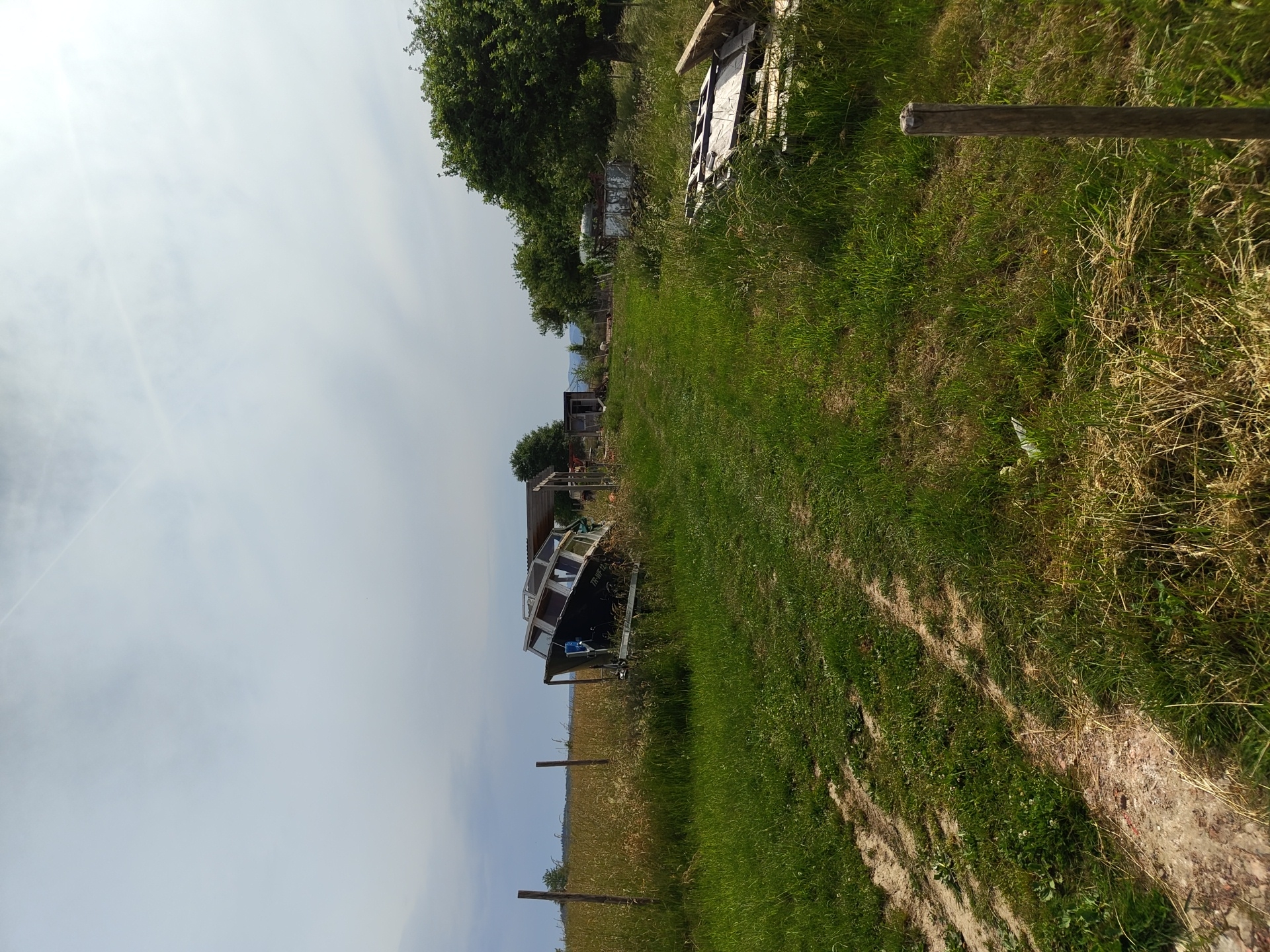 The image presents a rural or semi-rural scene, possibly located near Mettlach, Germany, based on the presence of typical Central European landscape elements. The foreground features a grassy area with a dirt track running through it. Patches of dry grass and weeds are interspersed with the green. A fence line with wooden posts runs along the left side of the frame, separating the grassy area from a field of brown, possibly harvested crops.
A small boat on a trailer is situated on the left side of the track, adjacent to the field. The boat appears to be in a state of disuse or storage. Behind the boat, there's a structure or shed. Further in the distance, more buildings and trees are visible. Piles of wood are positioned towards the rear right of the frame. The sky is overcast with a pale white-grey hue.