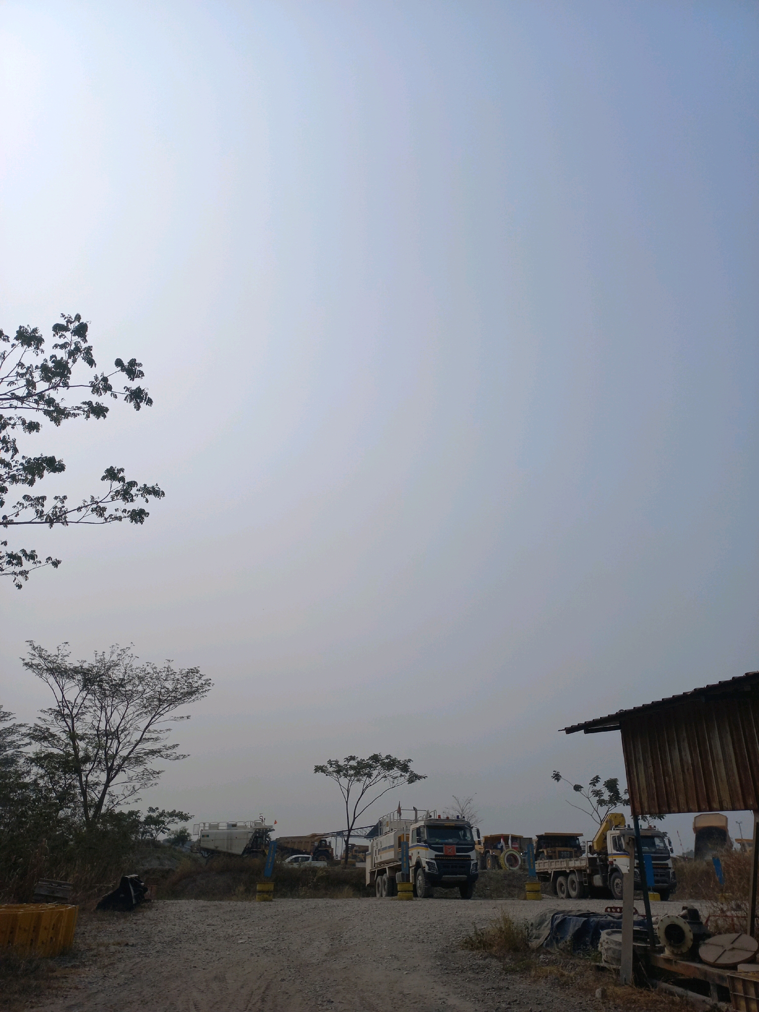 A wide shot captures a construction or industrial site in Paringin, Indonesia, under a hazy, light blue sky. In the foreground, a gravelly path leads towards several large vehicles, including dump trucks and excavators. The trucks are parked in a line, some with their beds raised. To the right, a rustic wooden structure with a corrugated metal roof stands partially in frame. A large tree with sparse foliage is visible on the left side, its branches reaching towards the sky. In the background, more trees and the outline of distant machinery can be seen. The overall scene is one of an active, outdoor work environment.