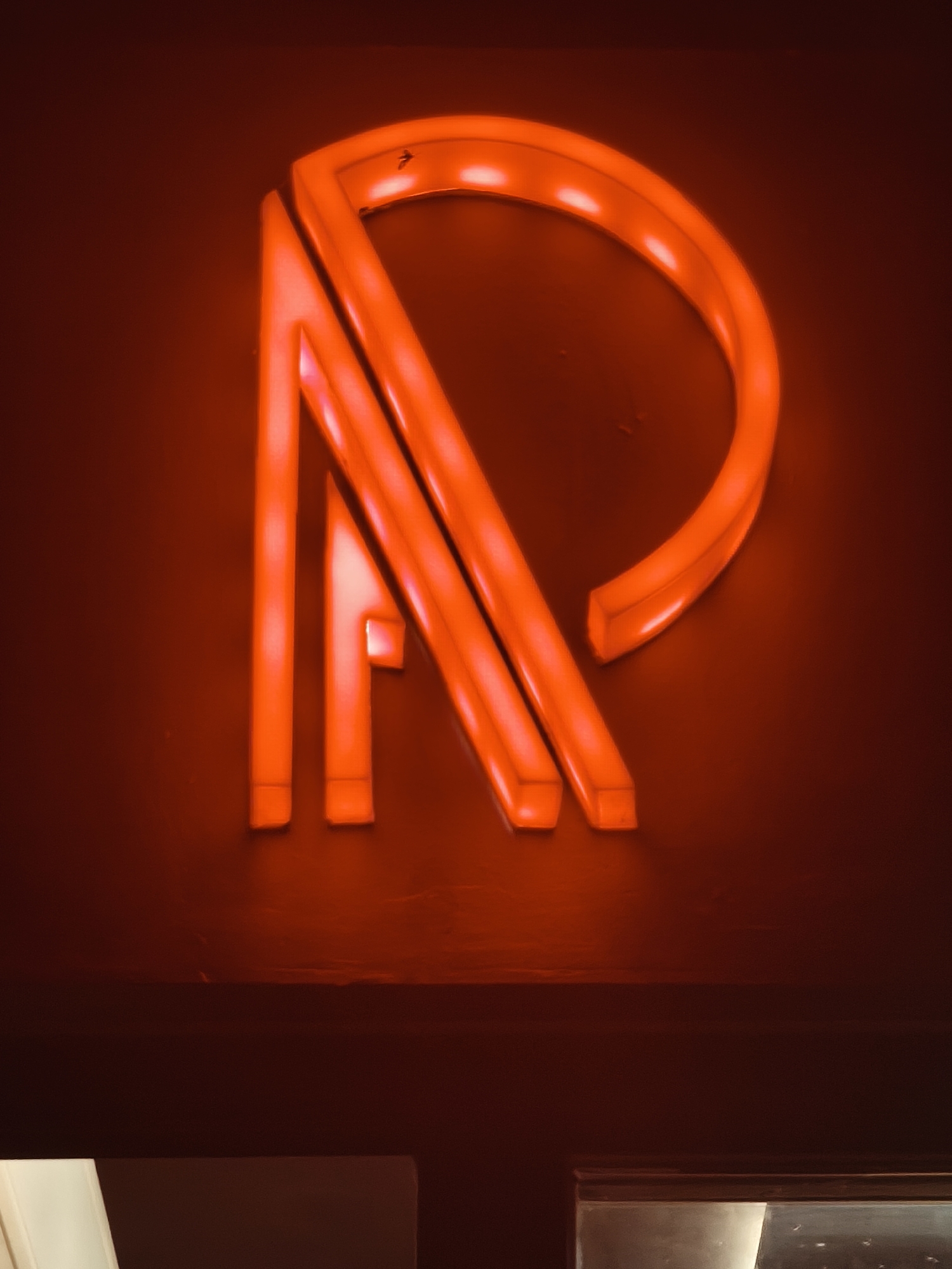 An abstract, red neon sign, likely depicting the letters "AP" or "RP", is illuminated against a dark, reddish-brown background. The sign consists of curved and angular neon tubes, forming a stylized design. The lower portion of the image shows a dark horizontal surface, possibly a shelf or ledge, and beneath that, a glimpse of a white object and a dark, reflective surface, suggesting part of a room or interior space. The overall impression is one of modern, possibly commercial signage. The location is identified as Kuta, Indonesia, though no specific landmarks or environmental details within Kuta are visible in the image.