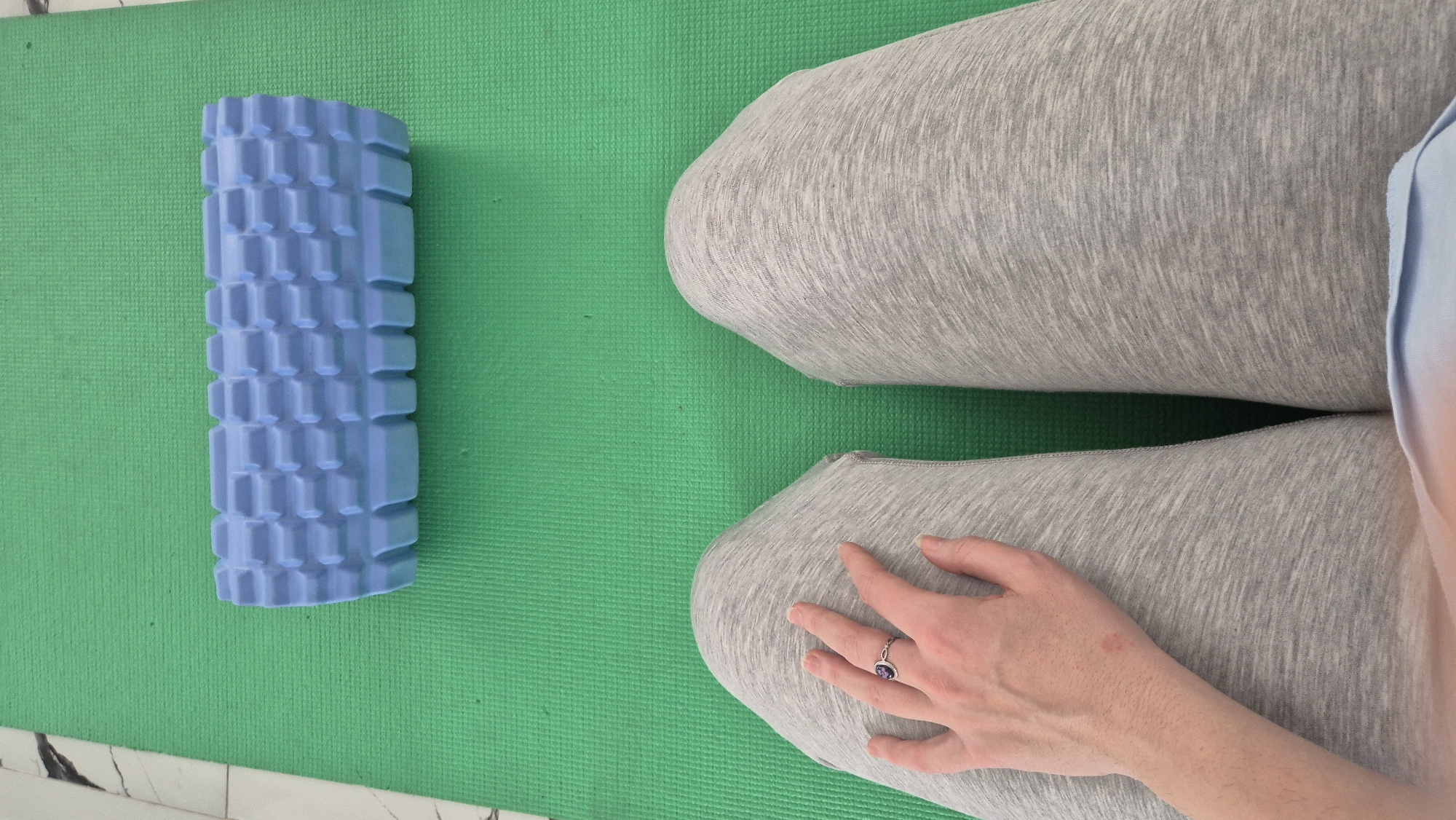 The image shows a green yoga mat on a white tiled floor. A blue foam roller sits to the left of a person wearing grey leggings, whose hand rests on their leg. The image provides no information to indicate that it was taken in Batumi, Georgia.