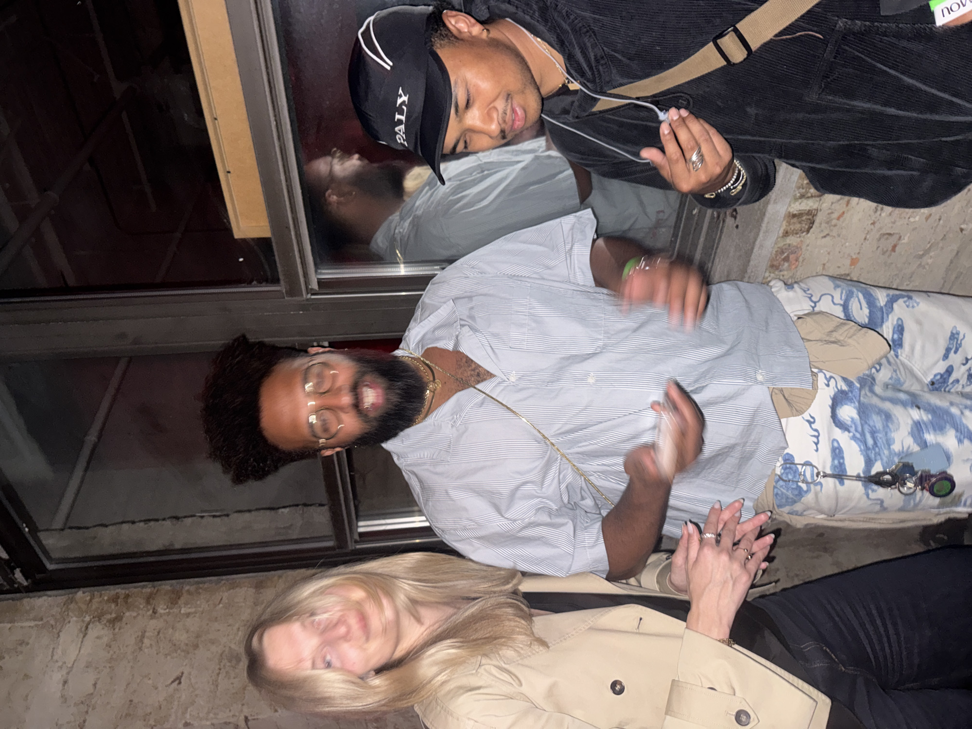 The image shows a group of three people standing near a window in New York City. One person is wearing a black corduroy jacket and a black baseball cap with "PALY" written on it. Another person is wearing glasses, a light blue and white striped shirt, and patterned pants. The third person has blonde hair and is wearing a beige trench coat and black pants. The reflection of the person in the black baseball cap can be seen in the window. The background suggests an urban setting.