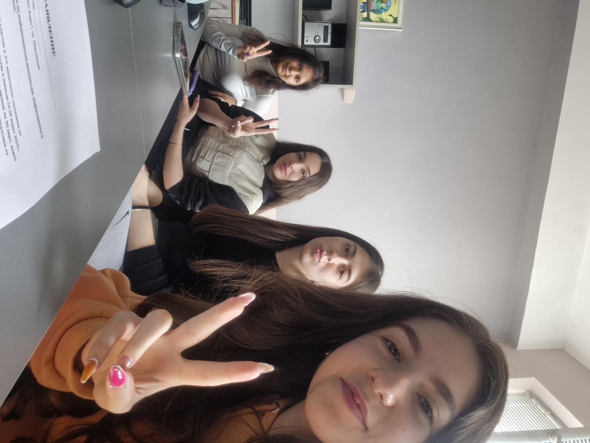 Four women are posing for a selfie. They are making peace signs with their hands. There is a document on the table in the lower left that states "Statement", and refers to participation in the 15th national competition in Pavlikeni, Bulgaria, from April 24-26, 2025.