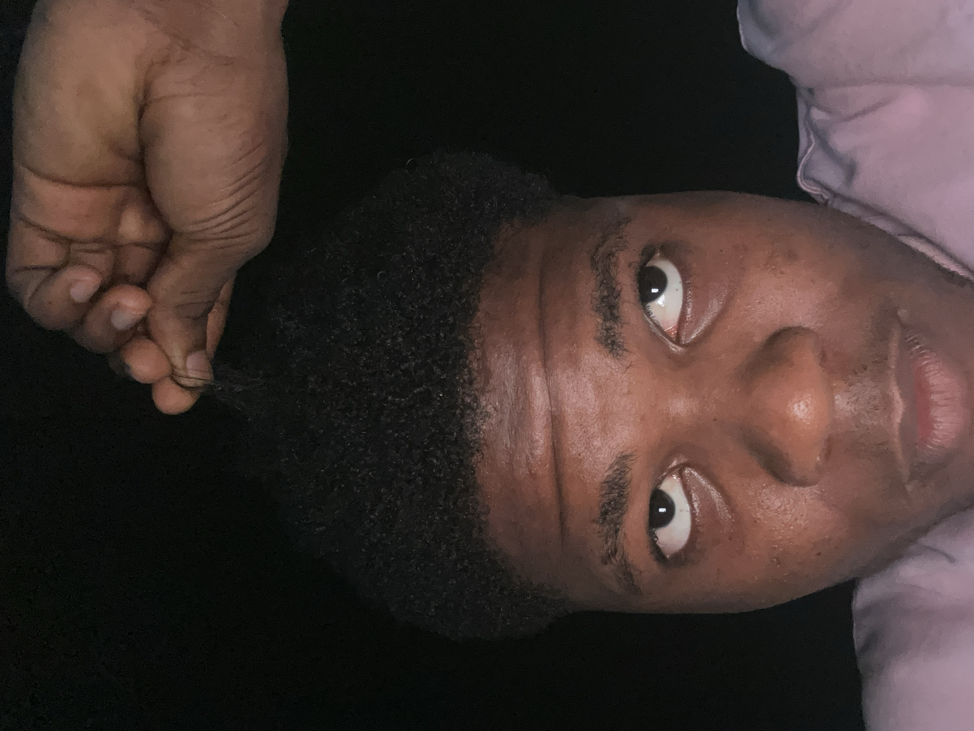 In a close-up, low-light photograph taken in Ihiagwa, Nigeria, a young Black male is depicted from the chest up. His dark, curly hair is styled short. His right hand is raised above his head, fingers grasping a few strands of hair. He is wearing a light pink garment. The background is entirely dark. The man's eyes are looking upwards and to his left. His facial skin exhibits some texture and minor blemishes.