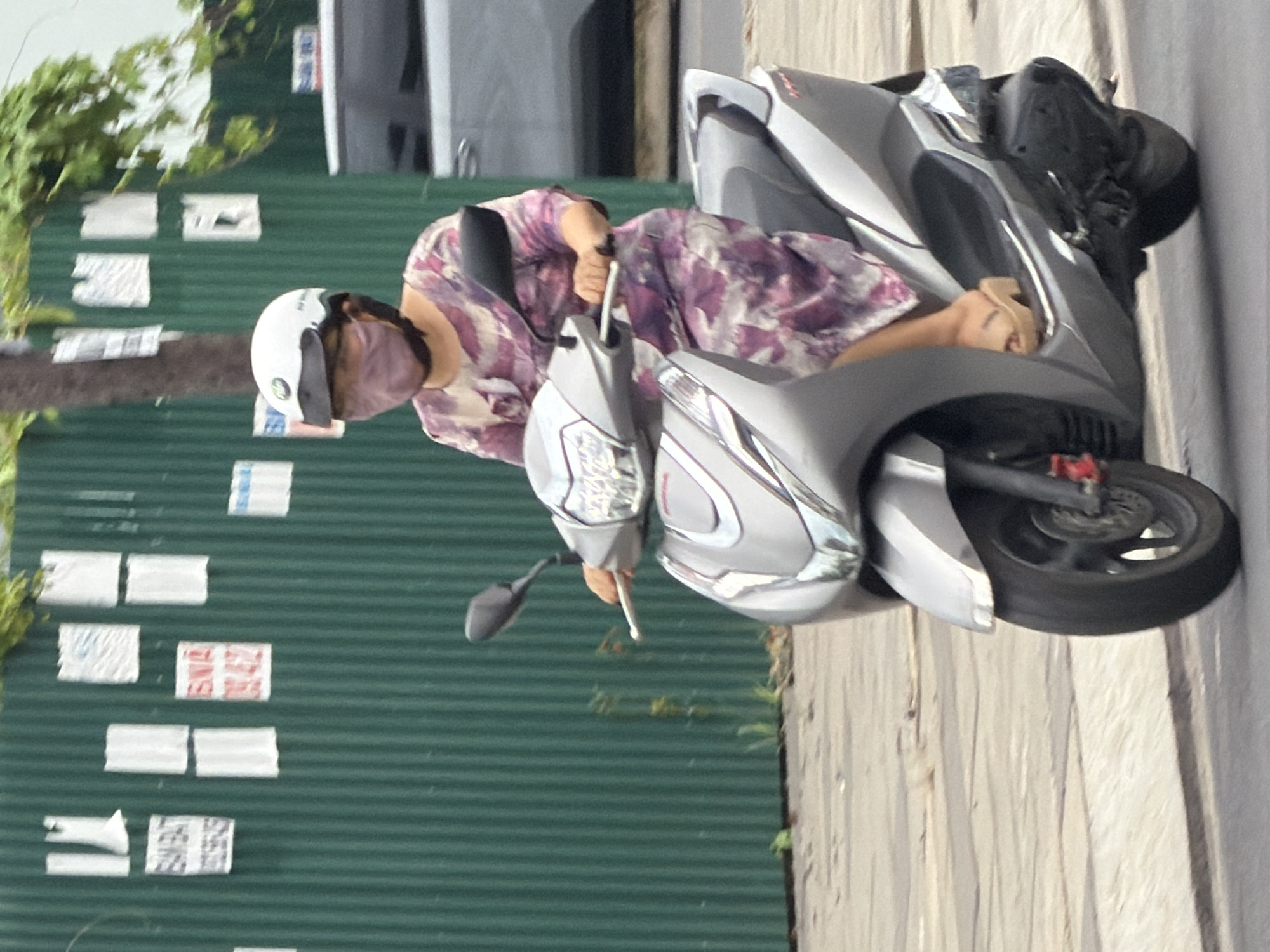 A person wearing a white helmet and a pink face mask rides a silver scooter in Nha Trang, Vietnam. The person is wearing a purple and pink floral dress and tan platform sandals. The scooter has a black seat and chrome accents. The background is a green corrugated metal fence with several posters attached to it. A tree with green leaves grows behind the scooter. The pavement in front of the scooter is a light gray concrete.