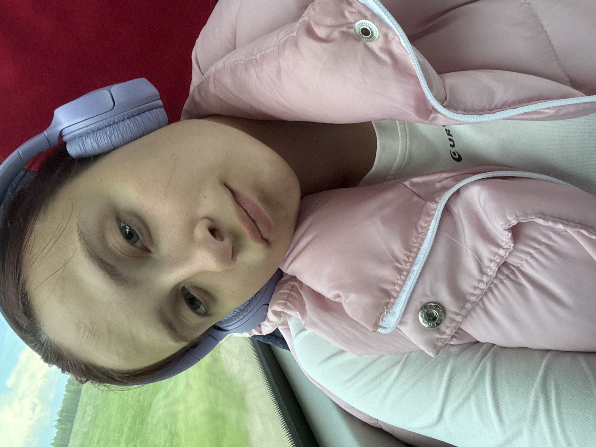 A young woman is pictured wearing lilac headphones and a pink puffer jacket, with a white t-shirt visible beneath the jacket. She appears to be inside a vehicle, possibly a train, as suggested by the blurred green landscape visible through a window in the background. There are no explicit cues in the image to confirm the location as Kukushtan, Russia.