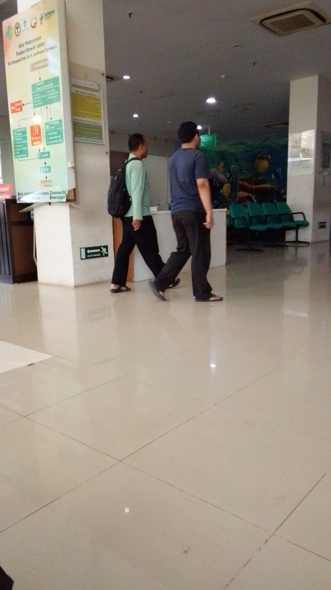 Two men are walking in a tiled hallway of what appears to be a hospital or public building in Pabelan, Indonesia. The man on the left is wearing a light green long-sleeved shirt, dark pants, and sandals, with a black backpack. The man on the right is wearing a dark blue t-shirt, dark pants, and sandals. They are walking towards the right side of the frame, away from the camera. To the left, there is a large informational poster with flowcharts and text. To the right, there is a waiting area with several green chairs and a mural depicting underwater life on the wall. The environment is well-lit with fluorescent lighting.