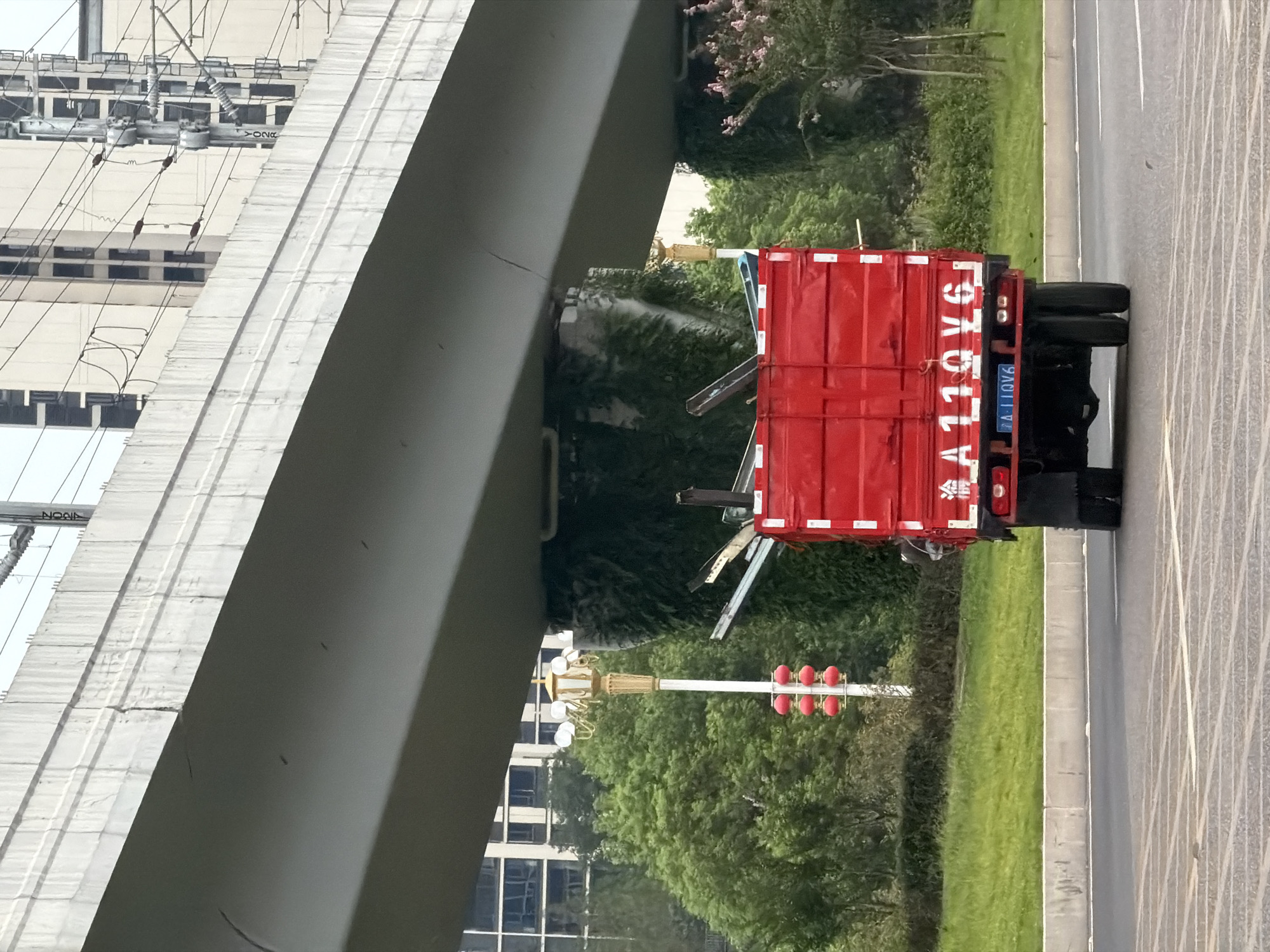 The image depicts a red flatbed truck driving on a road, partially obscured by an elevated concrete structure, likely a highway overpass or bridge. The truck's license plate is visible and reads "A 110V6". It is loaded with various metal poles or beams. In the background, lush green trees, buildings, and electrical infrastructure are present. A decorative lamp post with red lanterns stands to the left of the truck. The scene appears to be an urban or suburban environment in Jiangjin, China, indicated by the style of the truck, license plate, and the presence of Chinese characters on the truck. The overall lighting suggests it is daytime.