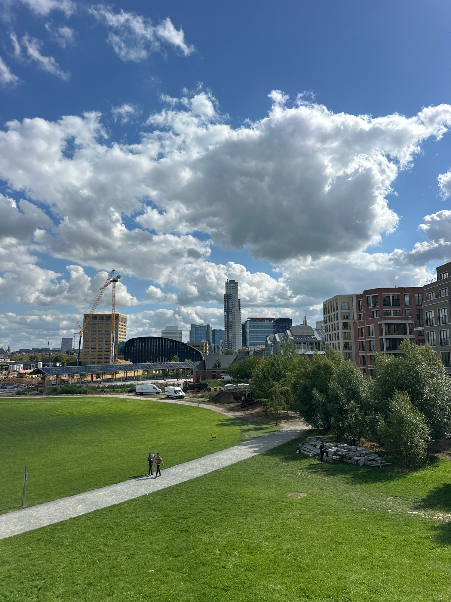 The image captures a sunlit day with dramatic clouds over a park area in Molenbeek-Saint-Jean - Sint-Jans-Molenbeek, Belgium. In the foreground, a well-maintained green lawn is bisected by a light-colored walking path. Two individuals are walking on the path, appearing to be strolling casually. To the right of the path, a pile of large, rough-hewn stone blocks is situated near a cluster of trees. Further into the midground, construction is evident with a tall yellow crane and various buildings under development. White vans are parked near a structure with solar panels. The background is dominated by a cityscape featuring a variety of modern buildings, including a tall, slender skyscraper and several multi-story residential and commercial structures. The sky is a vibrant blue, populated with voluminous, white cumulus clouds that cast shadows and create depth. The overall scene suggests a blend of urban development and recreational space.