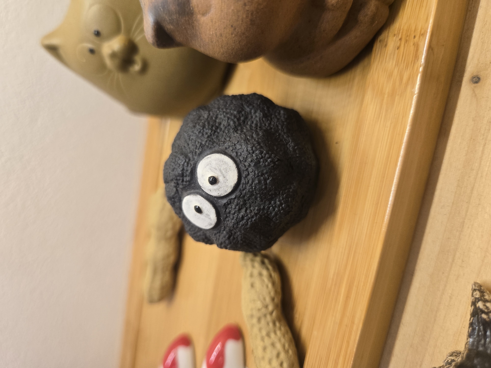 The image shows a close-up of a wooden surface with several decorative items attached. At the top left is a tan-colored ceramic cat-shaped object. Below it and slightly to the right is a small, round, black object with two white eyes, resembling a "soot sprite" character. Scattered around the sprite are several small, peanut-shaped objects. At the bottom left are two partially visible red and white striped objects. The overall composition suggests a display or collection of souvenirs or decorations mounted on a wooden backing. The location is not discernible from the image.