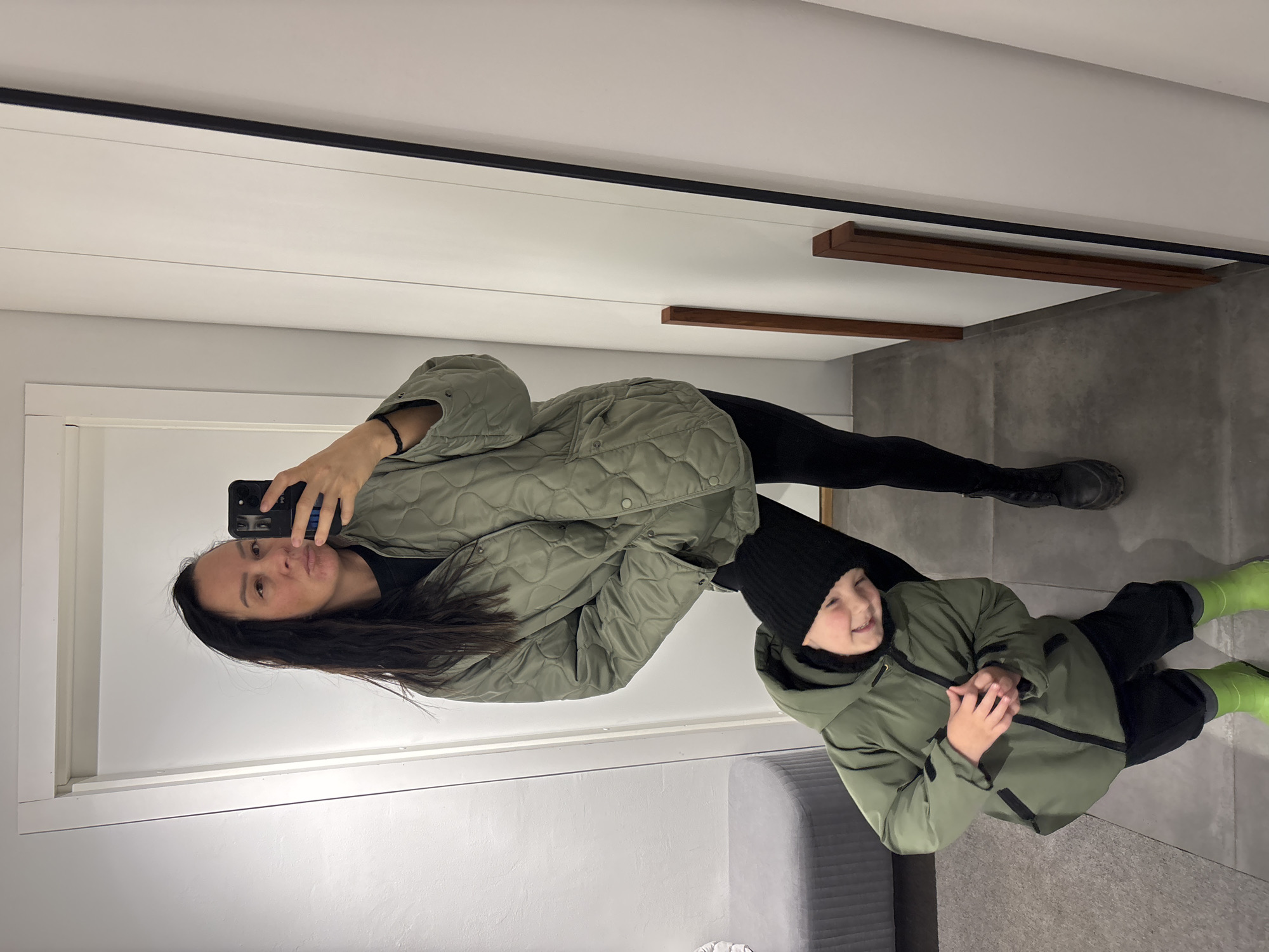 This mirror selfie, rotated to appear upright, captures a woman and a young child indoors in Chaikovsky, Russia.
The woman, positioned on the left, has long, dark hair and wears a quilted olive green jacket, black leggings, and black boots. She holds a black smartphone up, focusing on taking the picture, with a neutral expression. Her arm is adorned with a dark, braided bracelet.
Beside her, on the right, stands a young child, likely a boy, who is smiling widely. He is dressed in a black knit beanie, an olive green jacket similar in style to the woman's, black pants, and bright neon green boots. His hands are clasped together in front of him.
The indoor setting features white walls, a stark black horizontal trim running across the upper portion, and light grey tiled or concrete flooring. Decorative wooden panels or accents are visible horizontally on the far wall. A soft, grey upholstered piece of furniture, possibly a bench or seat, is partially visible in the lower right corner. The lighting is bright and uniform, suggesting an indoor artificial light source.
There is no visible text in the image. The scene conveys a casual, perhaps playful, moment between the two subjects, highlighted by their coordinated olive green jackets.