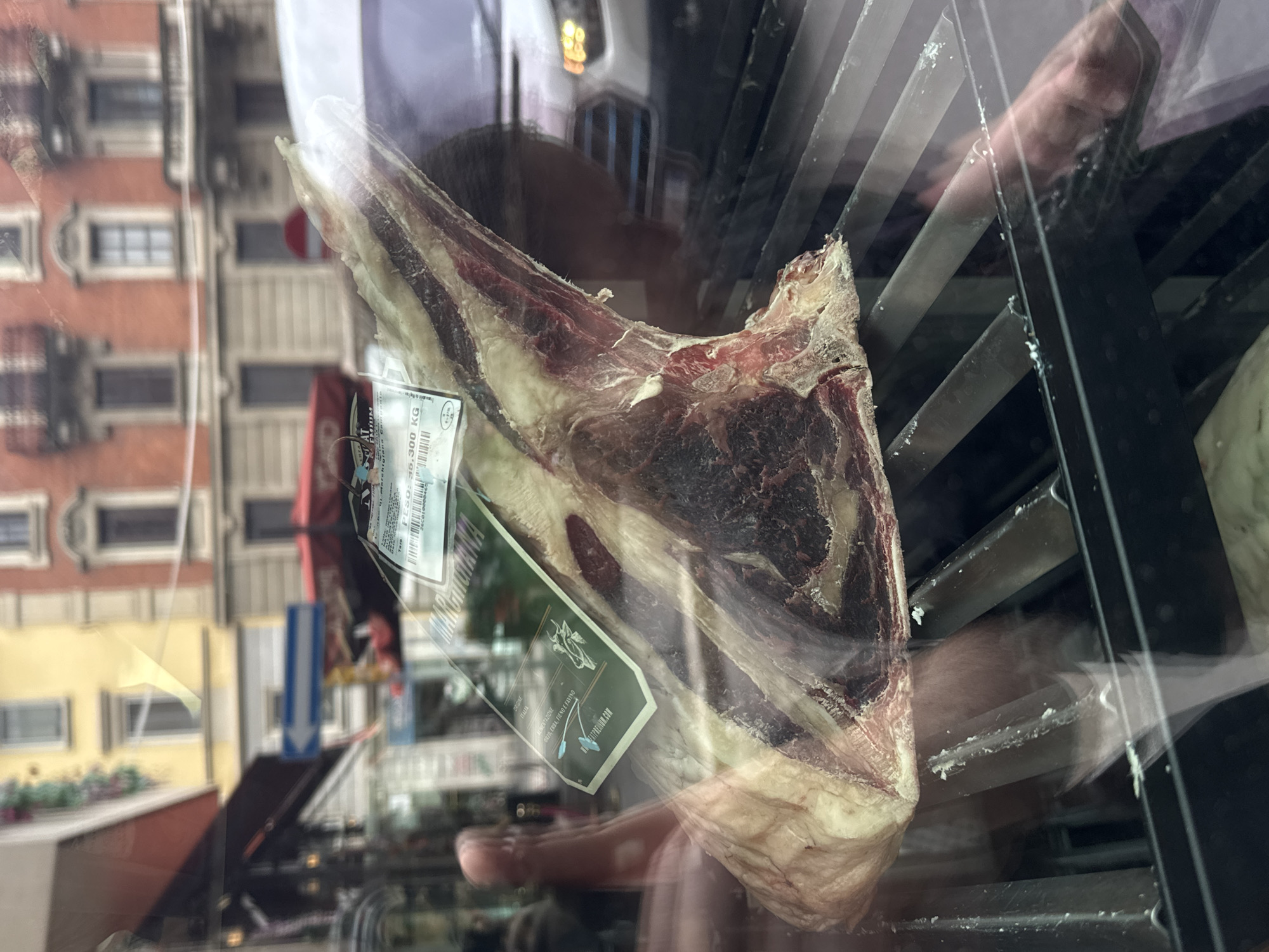 A large, raw cut of meat, likely beef, is displayed prominently behind a glass window. The meat, which appears to be a substantial bone-in primal cut, exhibits deep red muscle and significant white fat marbling, suggesting a high-quality product. It is partially encased in plastic or vacuum-sealed.
Attached to the meat are two labels. A green label prominently features the brand or type "MARANCIANA" and states its "ORIGINE ITALIA" (Origin: Italy). It also details the "ALIMENTAZIONE" (feeding) of the animal as "ORZO, ERBA, FIENO E FAVINO" (Barley, grass, hay, and broad beans), and includes a website, possibly "ATPREMIUM.COM", along with a logo of a stylized animal head. A white label provides a "PESO: 36,300 KG", indicating a substantial weight of 36.3 kilograms, and includes a barcode and the word "PREMIUM".
The glass display reflects the surrounding environment. Visible reflections include the distinct architecture of buildings with multiple windows, some in a reddish-brown brick and others in a lighter yellow hue, suggesting an urban street scene during daytime. A blue directional arrow sign is also reflected. A human hand, possibly belonging to the photographer, is clearly visible in the reflections, positioned towards the lower right of the meat. Through the glass, the interior of the display case shows metal racks or shelving, likely part of a refrigerated unit.
The scene depicts a butcher shop or specialty food store in Milan, Italy, showcasing a premium cut of meat for sale, with a passerby capturing the scene through the window.