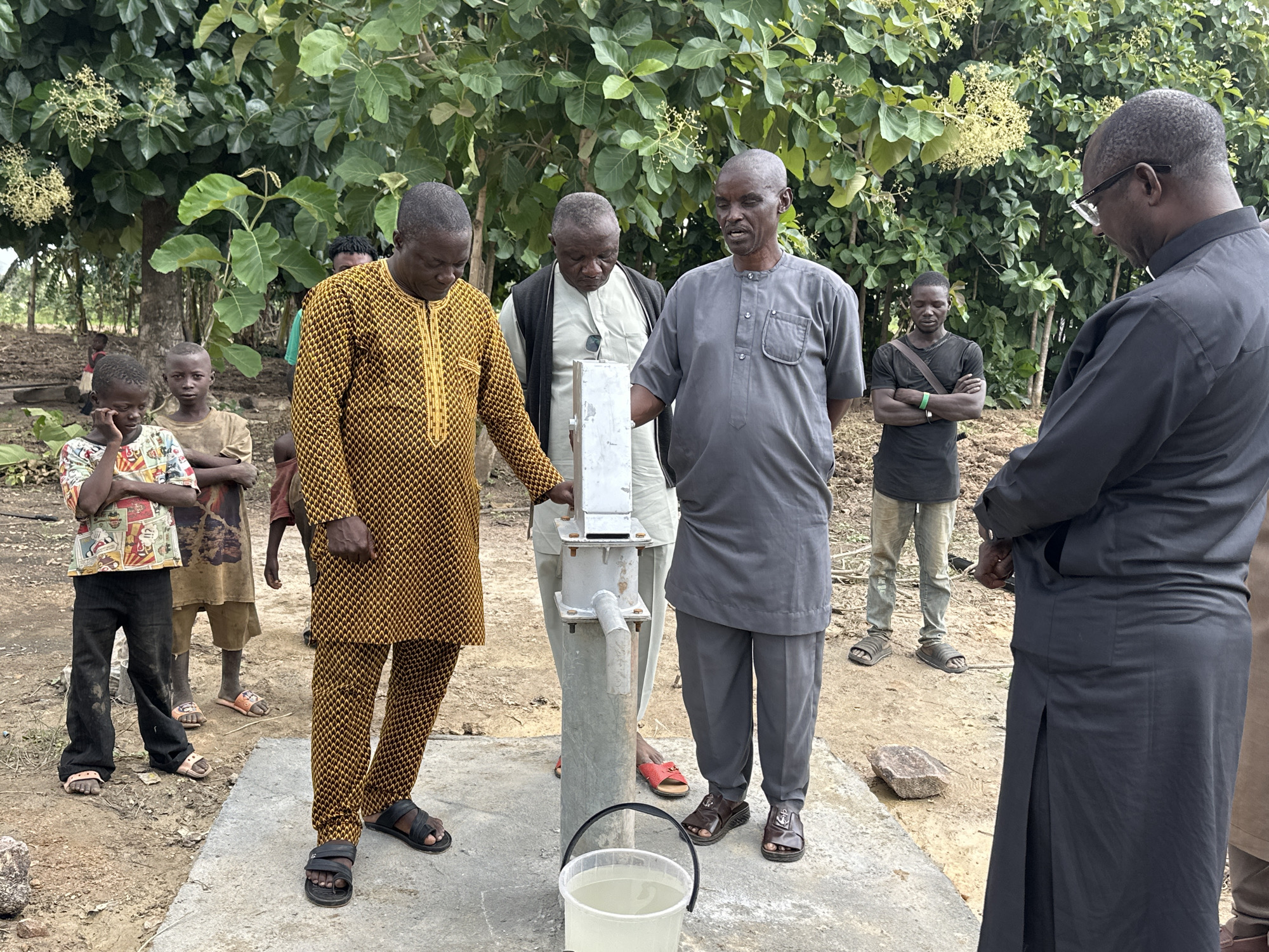 In Afogo, Nigeria, a group of seven individuals, including four men and three children, are gathered around a water pump. The men, dressed in traditional African attire and casual clothing, appear to be observing or operating the pump, which is situated on a concrete base. A white bucket filled with water sits at the base of the pump. The background consists of lush green foliage and trees, suggesting an outdoor, rural setting. The ground is covered in soil and scattered rocks.