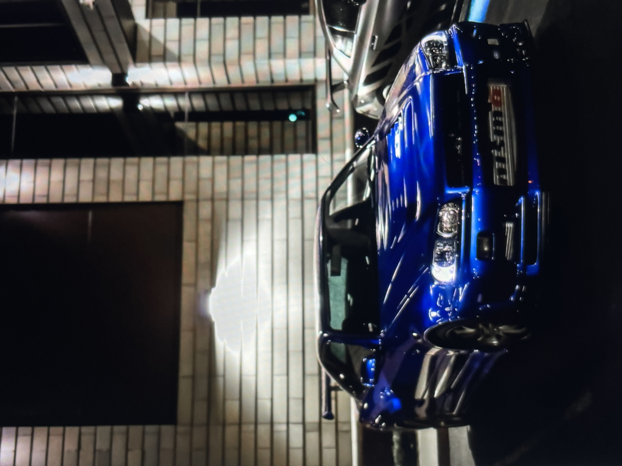 A blue Nissan Nismo car is parked next to a building in a city. The building has a brick facade and a large black window. The city is not specified. The image is taken at night.