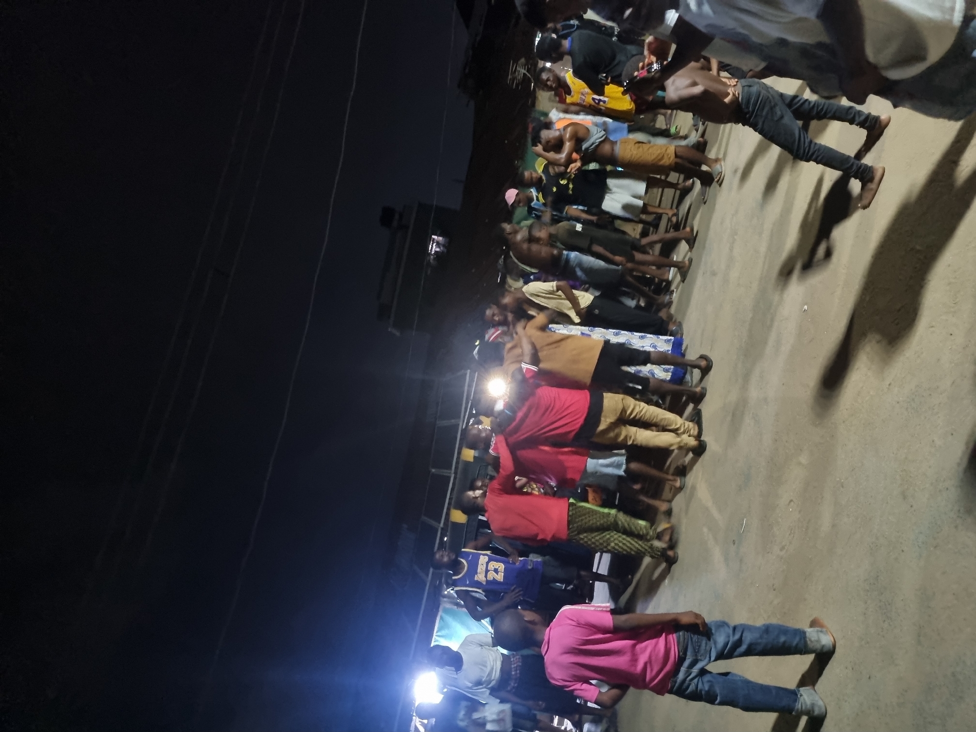 A nighttime scene shows a large group of people standing together outdoors in Onitsha, Nigeria. The group comprises men and young boys, some shirtless or wearing casual clothing like t-shirts and shorts. A few are wearing basketball jerseys, including one with the number 23 visible. They are standing on a paved surface, possibly a road or courtyard. There's a bright light source illuminating the group. Overhead, power lines crisscross the dark sky. A building is visible in the background.