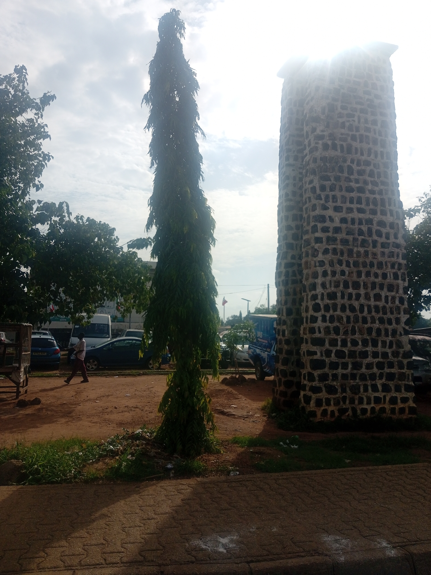 The image depicts an outdoor scene in Unguwar Rimi, Nigeria, under a bright, partly cloudy sky, suggesting it is daytime. The sun is intensely bright, causing lens flare and overexposure at the top right of the frame, partially obscuring a tall, stone tower. This tower, constructed from dark, rough-hewn stones of varying sizes, has a weathered, ancient appearance. To the left of the tower stands a very tall, slender tree with dense, feathery green foliage, reaching towards the sky.
In the middle ground, a person is walking from left to right across a dirt and gravel area. They are wearing dark trousers and a light-colored shirt. Several vehicles are parked or driving in the area, including a blue car on the left, a white van, and a blue and white patterned vehicle that appears to be a bus or truck. Lush green trees with full canopies are visible behind the vehicles and to the left, providing shade. The ground is a mixture of dirt, gravel, and patches of dry grass. A paved walkway with a textured pattern runs along the bottom of the frame. The overall atmosphere is one of a public space, possibly a street or intersection, with a mix of natural and man-made elements. There is no discernible text in the image.