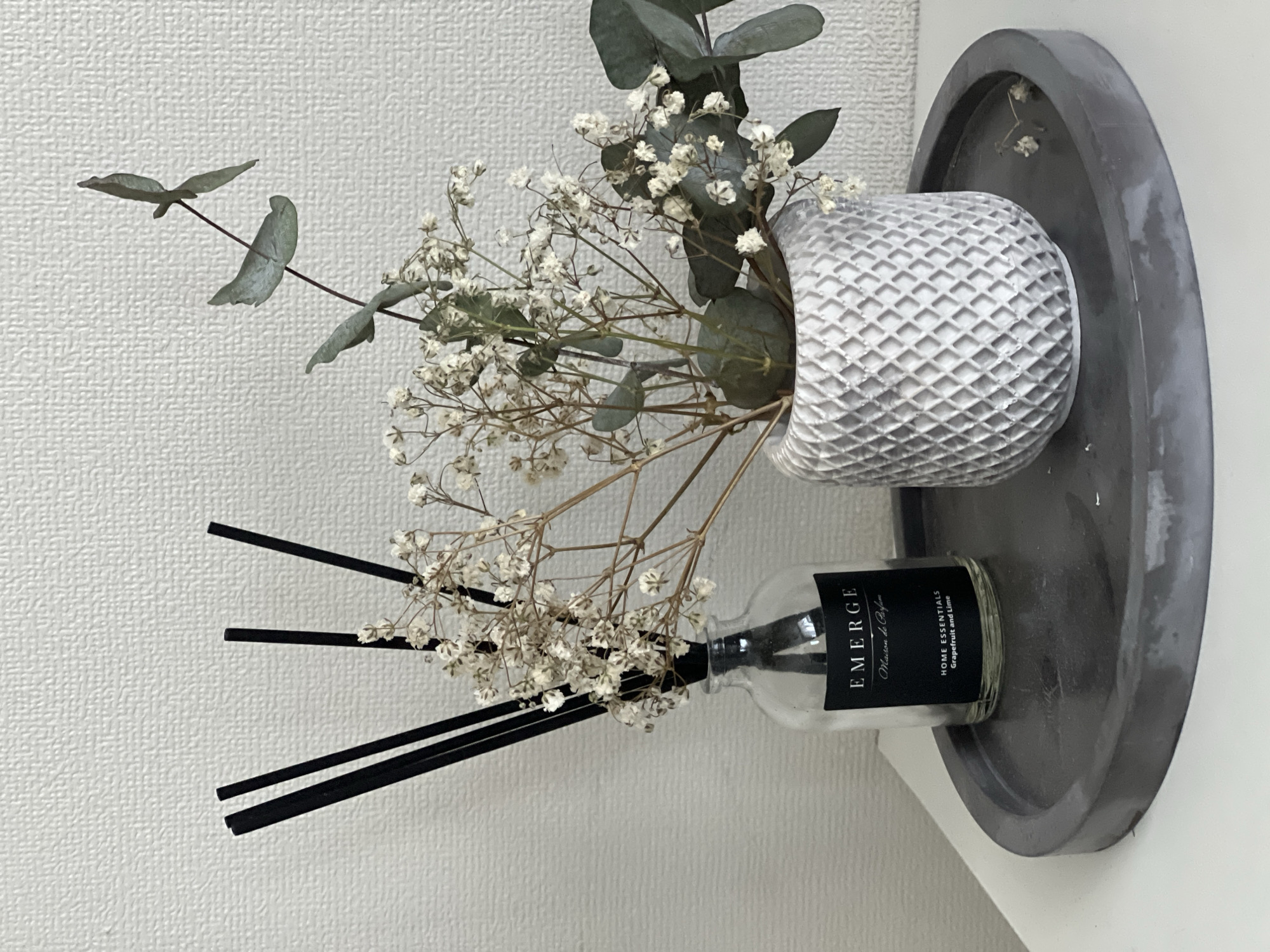 A gray decorative tray rests on a white surface. On the tray, a textured white vase holds baby's breath flowers and eucalyptus leaves. Beside the vase is a clear bottle with black diffuser sticks and a black label reading "EMERGE Maison de Parfum HOME ESSENTIALS Grapefruit and Lime." The background is a white textured wall. The location is Unknown, Unknown.