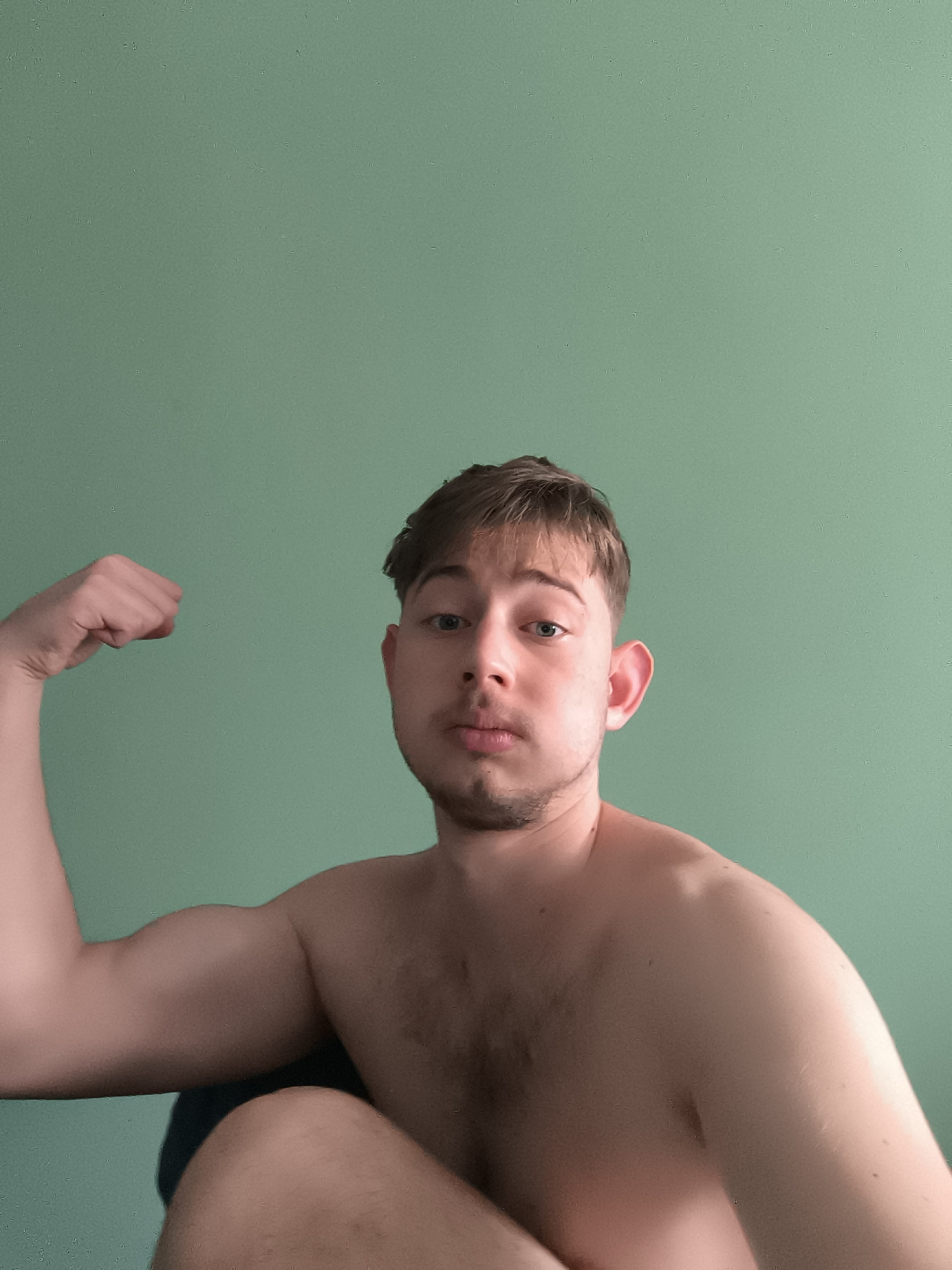 A shirtless, fair-skinned man with short brown hair flexes his left arm. He has light stubble and is looking directly at the camera. The background is a solid, light green wall. The location is unknown; there is no indication it is in Sofia, Bulgaria.