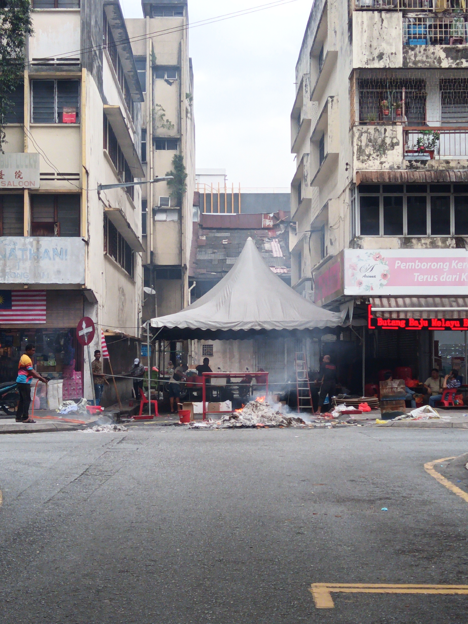 This is a street-level photograph taken in Kuala Lumpur, Malaysia, depicting an outdoor scene under an overcast sky. The image is dominated by several multi-story buildings with worn facades, characteristic of older urban architecture in the city. In the foreground, a street intersection is visible, with a patch of asphalt showing tire marks and some debris.
On the left side of the street, a man wearing a colorful shirt and dark pants is holding a hose, appearing to be watering something or cleaning the area. Beside him, a Malaysian flag is displayed on the facade of a building with a sign that reads "BUOY LUCKY SALOON".
The center of the image features a large white tent, likely for an outdoor event or market. Smoke is rising from a pile of burning material in front of the tent, creating a hazy atmosphere. Several individuals are present under and around the tent, some appearing to be vendors or participants in an activity. Red chairs and tables are visible within the tented area, as are various objects and containers.
To the right, another building facade is visible with signage. One sign reads "Pemborong Kere... Terus dari K...". Below that, a neon sign with red lettering says "Butang Baju Melayu". Several people are seated on chairs outside this establishment. The buildings exhibit signs of age, with peeling paint and visible structural elements. The overall impression is one of a bustling, albeit somewhat gritty, urban street scene in Kuala Lumpur.
