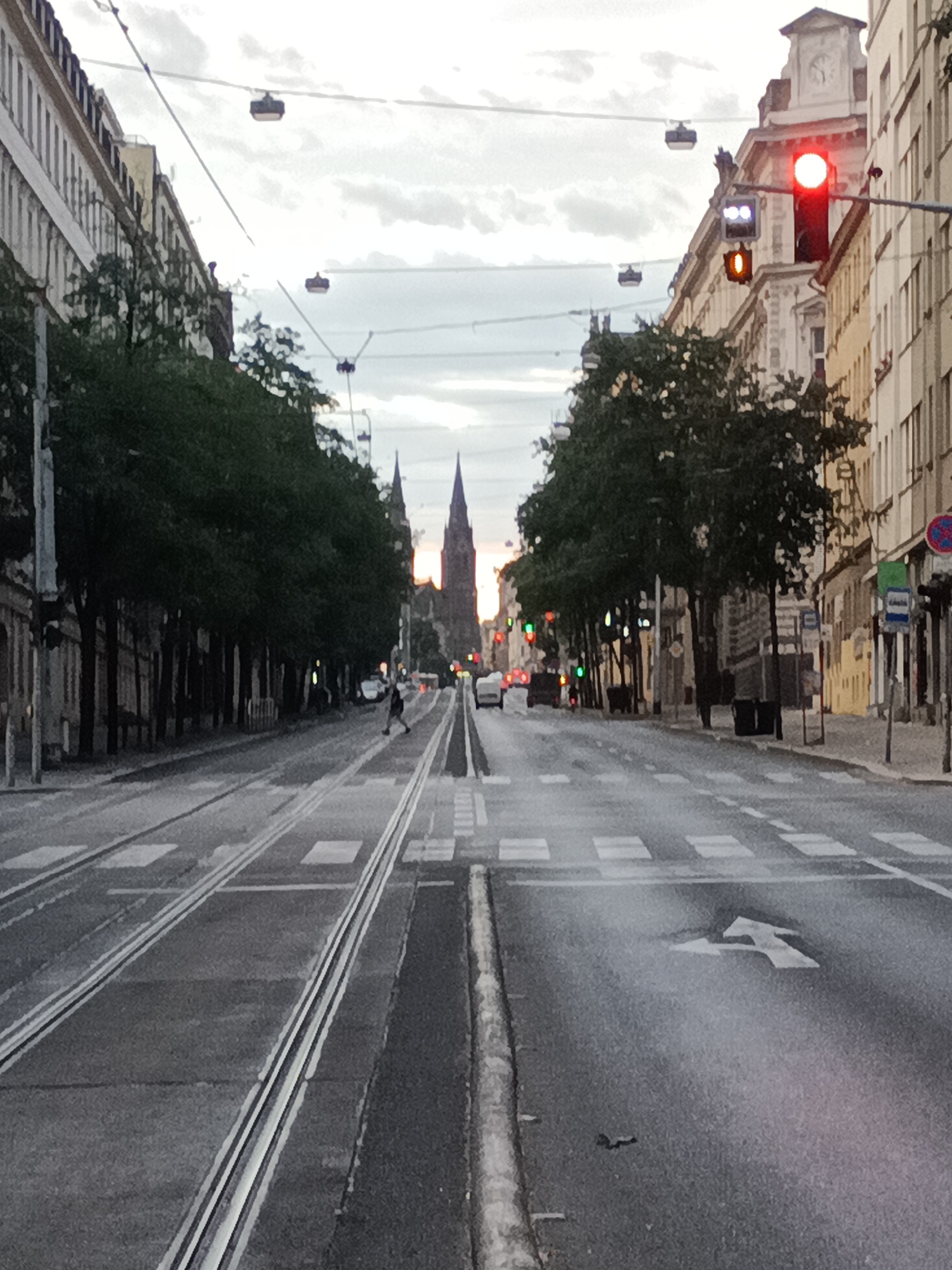 The image captures a street scene in Prague, Czechia, looking down a wide road with tram tracks. The sky is overcast. On the right side of the street, a building with light-colored facades and several windows is visible. A traffic light at an intersection shows a red light. Further down the street, the spires of a large, gothic-style church or cathedral can be seen. On the left side of the street, there are buildings with darker facades and trees lining the sidewalk. A person is crossing the street in the distance. In the foreground, the asphalt road is marked with pedestrian crossings and a left-turn arrow. The tram tracks are prominent in the center of the road. Overhead, wires for the tram system are strung across the street, with what appear to be attached housings.