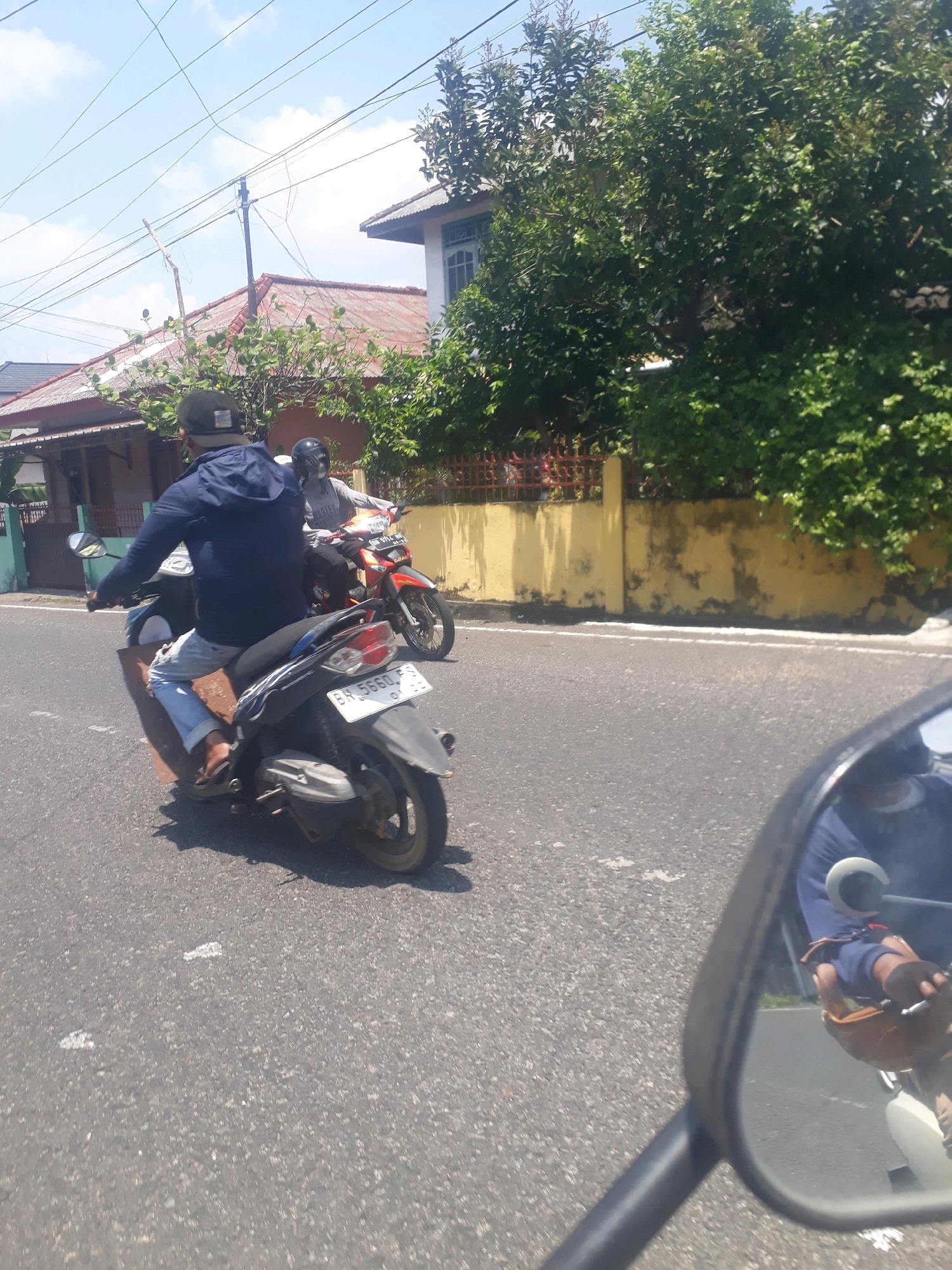 Two individuals are riding motorcycles on a paved road in Pangkalpinang, Indonesia. The primary subject is a person wearing a dark blue jacket and a black baseball cap, riding a black scooter. This individual is seen from behind, with their right leg extended forward. A second person, wearing a light gray shirt and a helmet, is on an orange and black motorcycle to the right and slightly ahead of the primary subject. The motorcycles have visible license plates, suggesting they are legally operated.
The environment includes residential buildings with red-tiled roofs and painted walls on the left side of the road. Lush green trees and foliage are visible, contributing to a tropical atmosphere. A yellow wall runs along the right side of the road, behind the second motorcycle, with some signs of wear and tear. The sky is bright blue with scattered white clouds. The scene appears to be illuminated by strong sunlight, casting shadows. The presence of utility poles and overhead wires indicates an urban or semi-urban setting. The image seems to be taken from the perspective of another vehicle, possibly a third motorcycle, as indicated by the reflection of the rider and a mirror in the foreground.