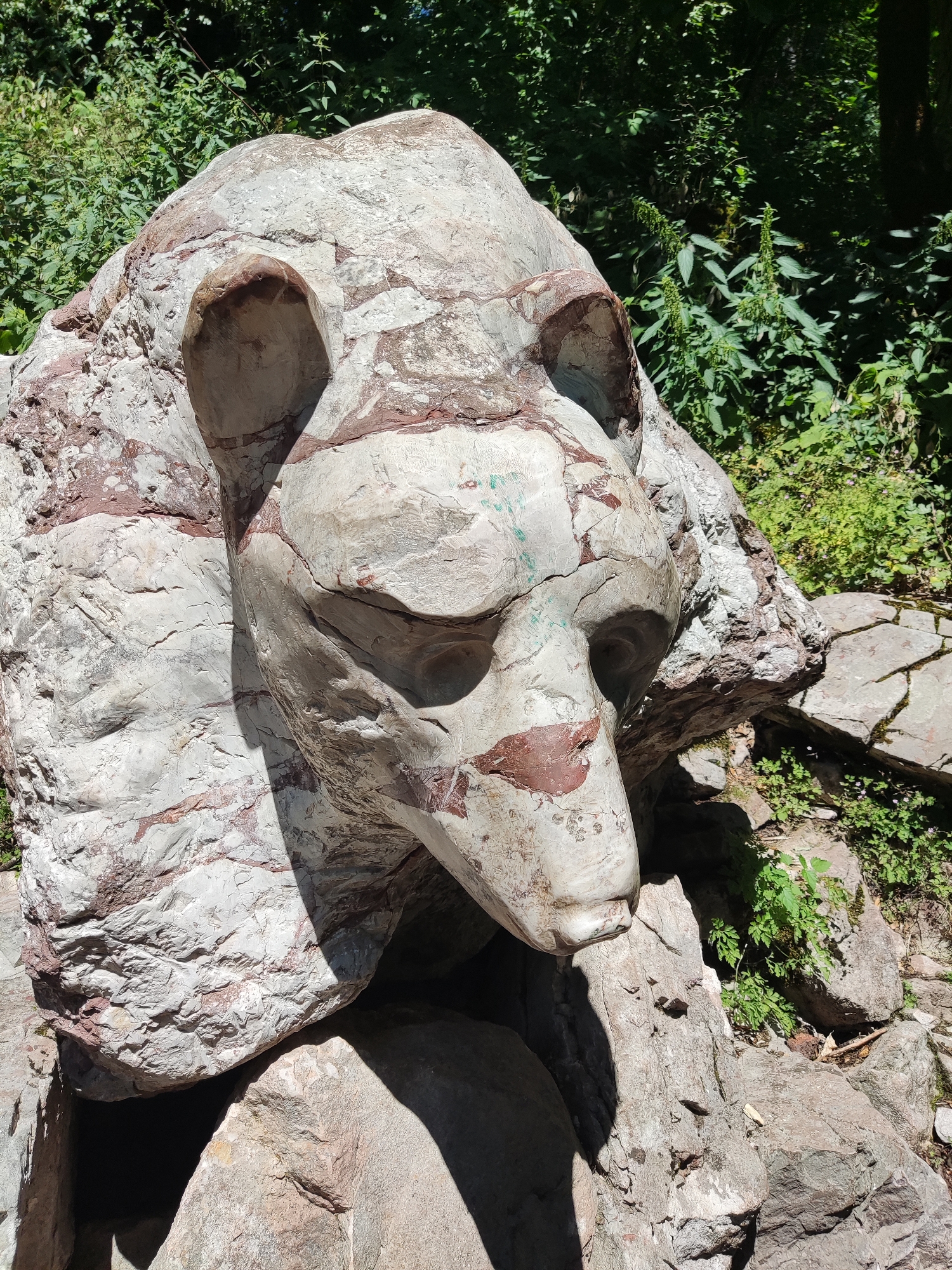 The image shows a large rock formation carved into the head of a bear. The bear's head is carved from a light-colored, veined rock with reddish-brown markings. The bear's eyes are deep-set, and its snout protrudes forward. The rock is situated outdoors, surrounded by lush green vegetation, suggesting a natural or park-like setting. The lighting indicates it is daytime, with shadows cast across the bear's face and the surrounding rocks. The specific location within Sjerogošte, Montenegro is not discernible from the image itself. There are no people or animals present in the image.