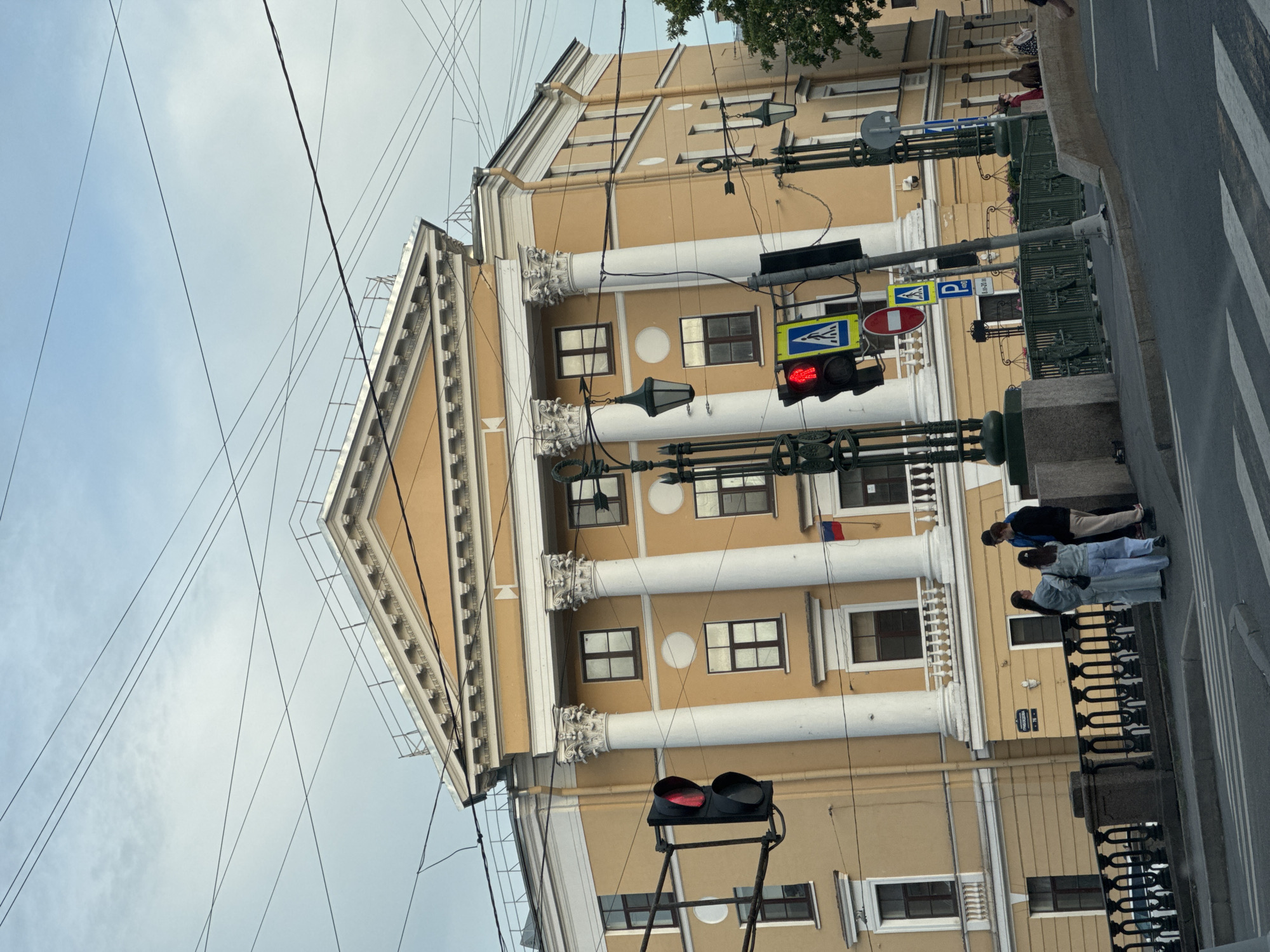 The image captures a street scene in Saint Petersburg, Russia, dominated by a large, neoclassical building with a yellow facade, prominent white columns, and an ornate pediment. Several power lines crisscross the sky above. In the foreground, a pedestrian crossing is visible, along with a traffic light showing red, and two blue pedestrian crossing signs. A group of four individuals, two women in light-colored outfits and two men, stand on the sidewalk near a decorative black metal fence. To the right, a green bridge railing and more traffic signage are present, including a parking sign. The overall environment suggests an urban setting in a historic district.