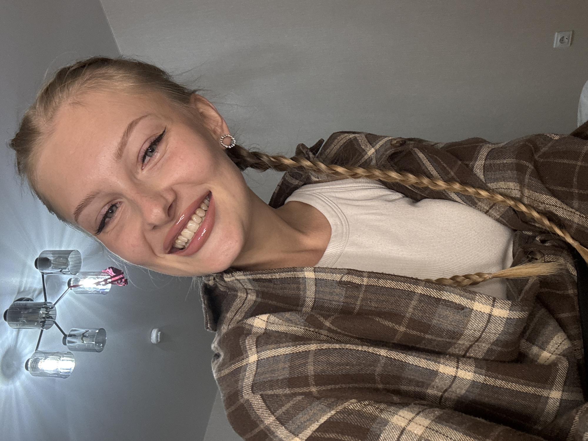 An unidentifiable female subject, with blonde hair in two braids and a large smile, is centered in the frame of the image. She is wearing a tan and brown plaid flannel shirt over a white ribbed tank top. A modern chandelier with glass shades hangs from the ceiling in the upper left corner of the image. The background consists of a plain gray wall with a light switch and a power outlet visible on the right. There is no discernible urban or city context provided.