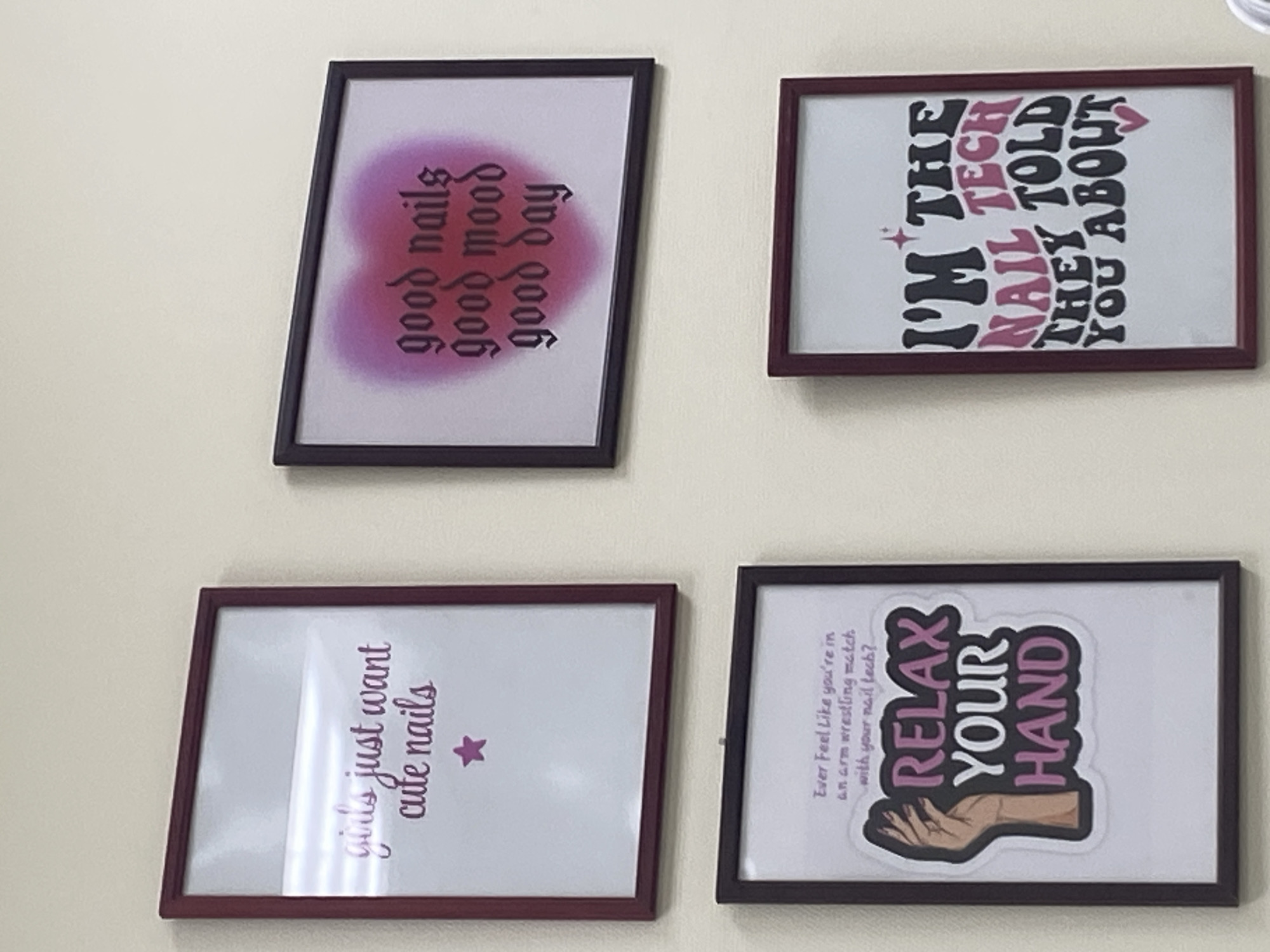 The image shows four framed prints hanging on a light-colored wall. From top left to bottom right, the prints read: "girls just want cute nails" with a pink star; "good nails good mood good day" in gothic script with a blurred pink heart background; "Ever feel like you're in an arm wrestling match with your nail tech? RELAX YOUR HAND" with an illustration of a hand; and "I'M THE NAIL TECH THEY TOLD YOU ABOUT" in stylized lettering with pink and black colors and small heart and star accents. The frames are dark brown and burgundy. The scene appears to be indoors, likely a nail salon or a similar business, and no people are visible. The location is not Kazan, Russia.