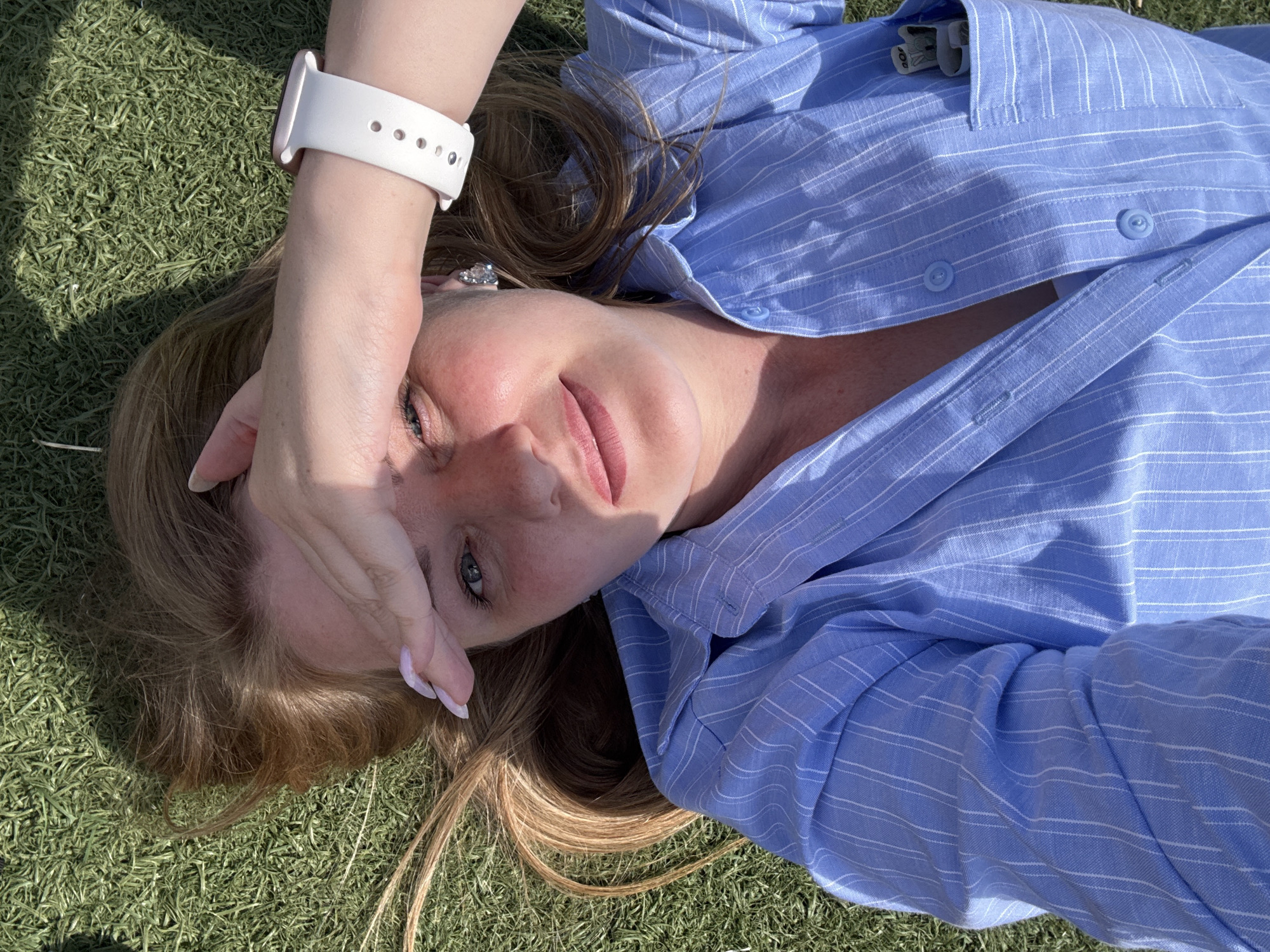This is a close-up, overhead shot of a woman lying on artificial grass. She is wearing a light blue, pinstriped button-down shirt, and a white smartwatch on her left wrist. Her right hand is raised, covering part of her face, with manicured fingernails visible. The woman has blonde hair that is spread out around her head. Sunlight is casting shadows on the grass and on her face, suggesting it is daytime. The overall mood appears relaxed and casual. The image does not contain any visible text. The location context provided indicates Chaikovsky, Russia, but there are no specific visual cues in the image to confirm this.