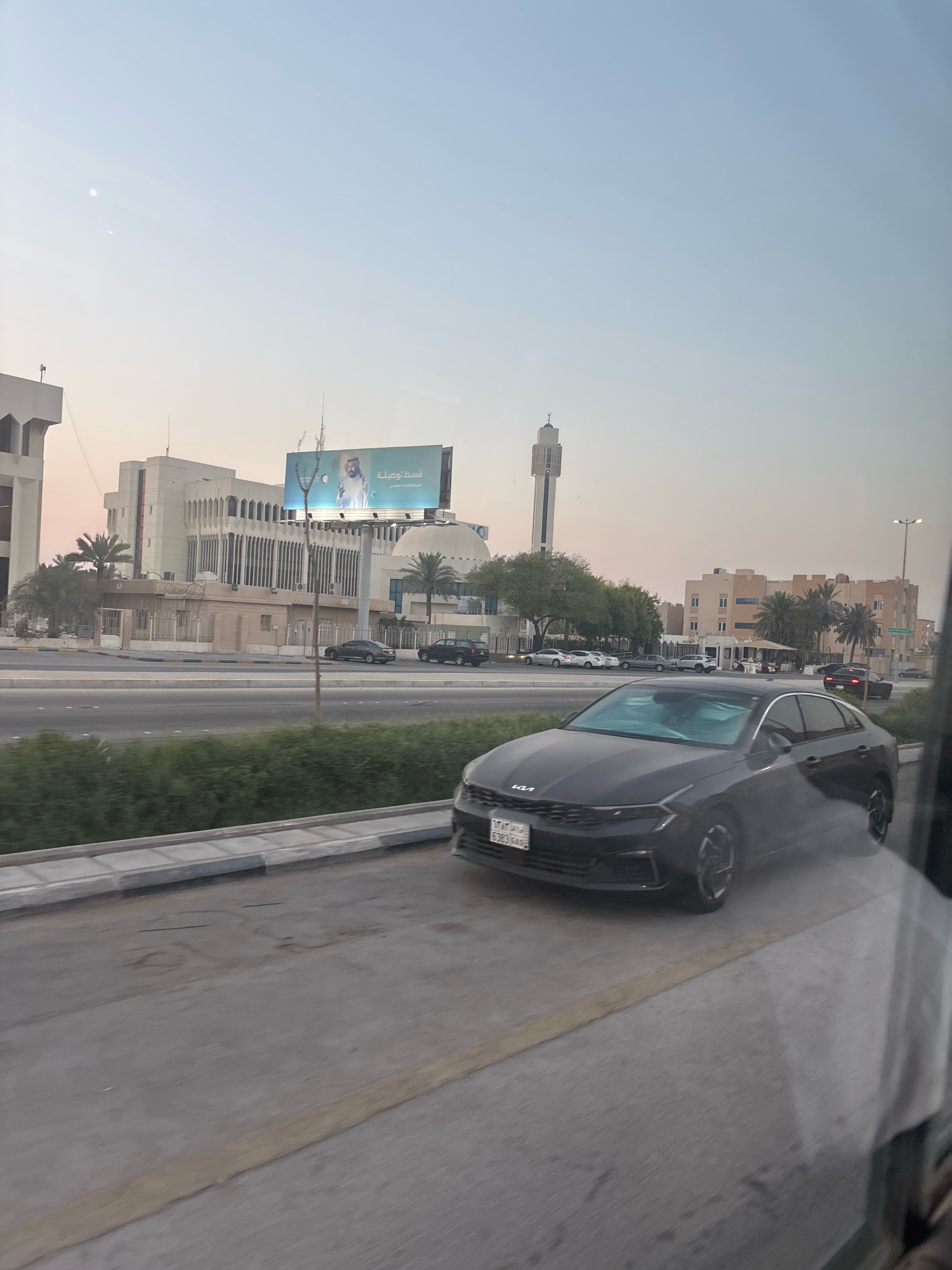 In Dhahran, Saudi Arabia, on a bright, clear day, a dark-colored Kia sedan drives along an asphalt road, its front license plate displaying Arabic script and numbers. The car is positioned in the foreground, capturing a significant portion of the frame. Beyond the road, a median of green bushes separates the traffic lanes. In the background, several buildings are visible, including a modern mosque with a tall minaret, and an office building with a distinctive architectural style characterized by vertical lines. A large billboard displaying an image of a man in traditional Saudi attire and Arabic text is mounted on a structure between the buildings. Other vehicles, including dark sedans and SUVs, are seen on the road in the middle ground. Palm trees and other greenery dot the landscape, suggesting a warm climate. The sky above is a pale blue, with a hint of haze. The scene is captured from a slightly elevated perspective, possibly from a moving vehicle, indicated by the slight blur and reflection on the right side of the image.