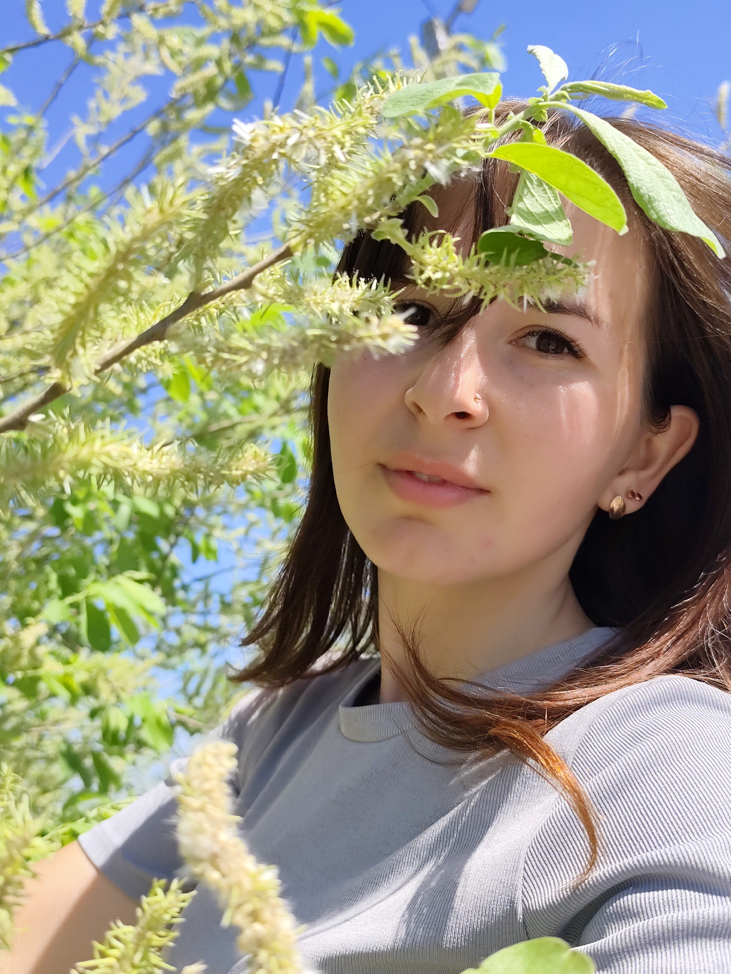 A young woman with brown hair stands outdoors, partially obscured by branches of a flowering tree with green leaves and light-colored catkins. The background reveals a blue sky and more green foliage, suggesting a natural setting. She wears a light gray top and gold earrings, with a small stud visible in her nose. She is looking directly at the camera. There is no information available about the location in the city of Фоки, Russia, from the image.