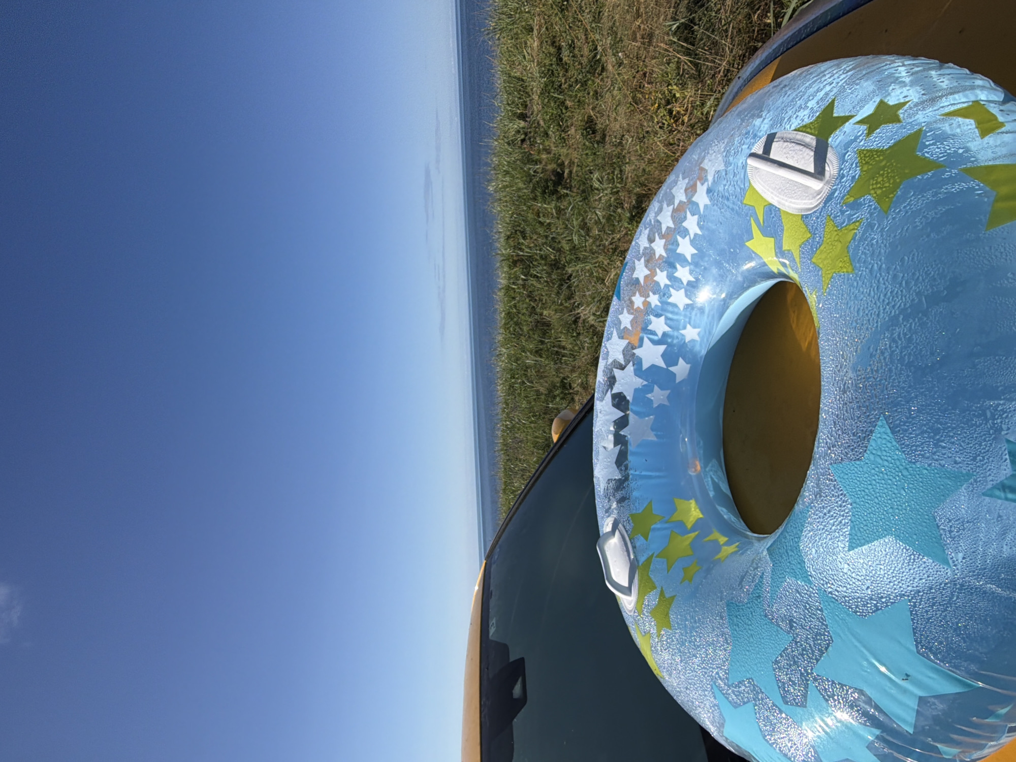 An inflatable blue and yellow star-patterned inner tube is resting on the hood of a yellow car, parked on a grassy embankment overlooking the sea. The sky is clear blue with a single small cloud. The water is calm and stretches to the horizon. The inner tube appears slightly wet, possibly from dew or sea spray. The car's windshield and part of the dashboard are visible. The scene is devoid of people. The location is Ozerskoe, Russia.
