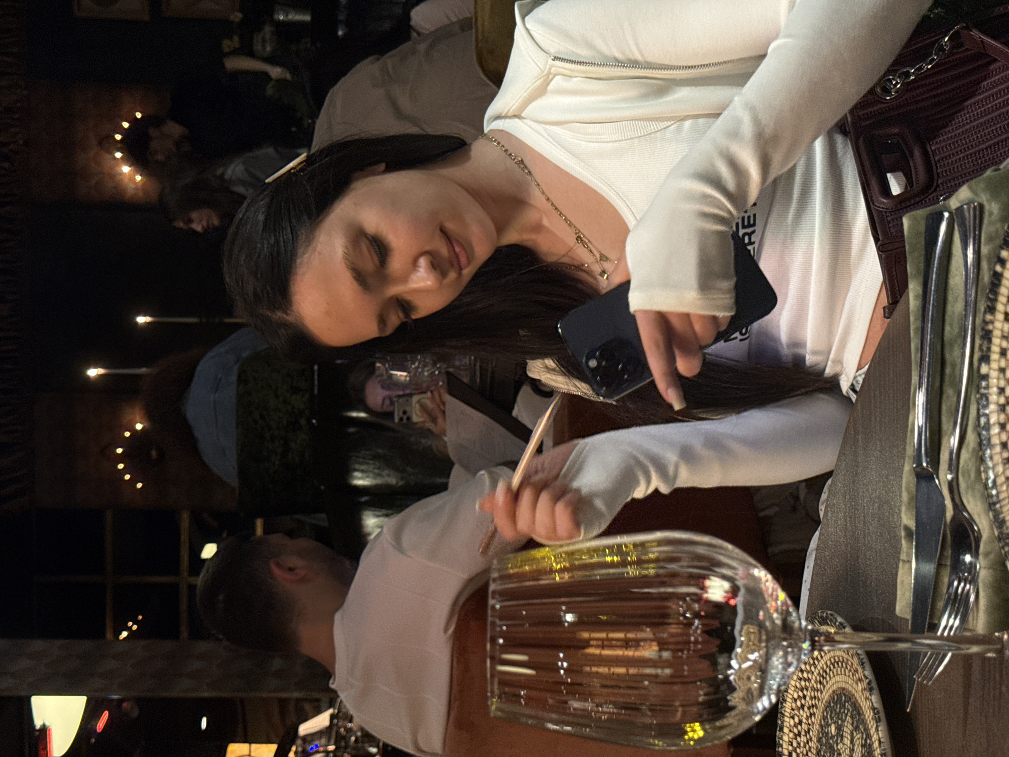 The scene appears to be inside a restaurant or bar. A woman with long dark hair is seated at a table, holding a smartphone in one hand and a thin rod, possibly a stir stick or similar object, in the other. She is wearing a white top and a necklace. Next to her, on the table, is a clear glass, likely a wine glass, and cutlery. A man in a white shirt is partially visible on the left side of the frame. Other individuals and lighting are visible in the background.
While the city of Yerevan, Armenia cannot be confirmed with certainty from the information in the image, the scene depicts an indoor dining environment and could potentially be located there.