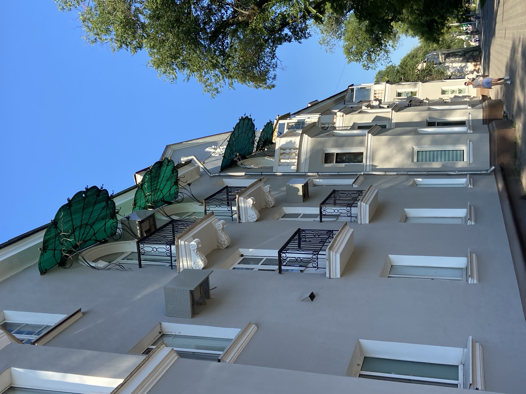 The image shows a close-up, low-angle view of a light grey building with ornate white trim around its windows and balconies. Several balconies are visible, each with black metal railings and decorative white supports. Green mesh awnings are attached to the upper balconies. An air conditioning unit is mounted on the side of the building. The sky is clear and blue, and a large green tree is visible on the right side of the frame. The building appears to be on a street in Blagoveshchensk, Russia, as indicated by the context provided. In the background, a woman is walking away from the camera on a paved walkway lined with trees.