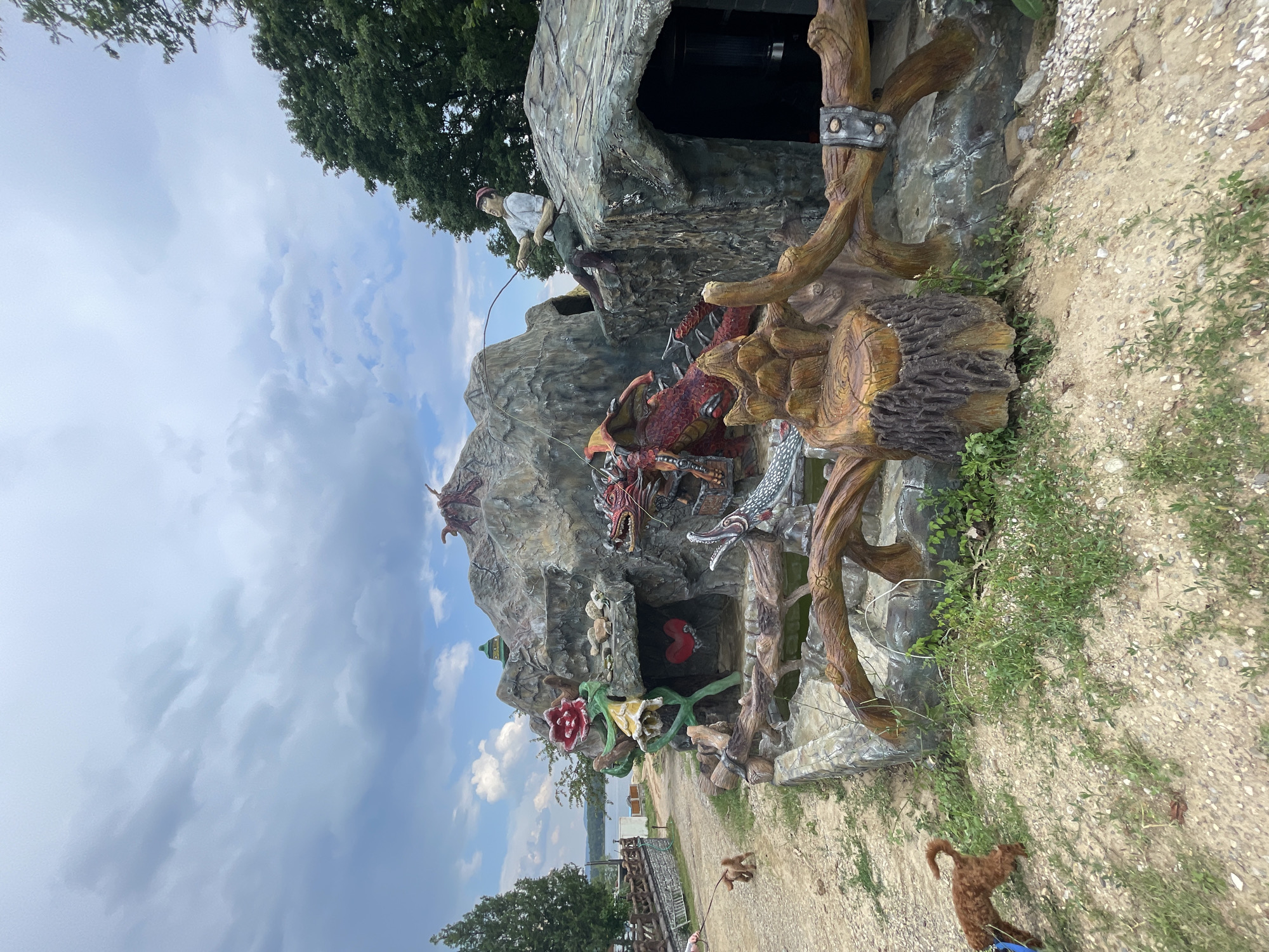This image depicts an outdoor art installation in Koprinka, Bulgaria, set against a natural landscape. The central feature is a large, multi-level artificial rock formation constructed from concrete or similar material, painted in natural tones.
On this rock formation, several statues are integrated:
- A prominent red dragon with dark wings and sharp teeth is positioned on the lower right, appearing to emerge from a cave-like opening.
- Above the dragon, on the highest peak of the formation, a smaller, reddish, multi-limbed creature is visible.
- Below the red dragon, a white or silver serpent-like creature with an open mouth is present, partially submerged in a trough.
- A stylized red rose sculpture, featuring green stems and leaves, is affixed to the upper-left section of the rock.
- Beneath the red rose, a yellow, bell-shaped flower sculpture is attached.
- A red heart sculpture is visible within a dark, cave-like recess below the yellow flower.
- On the upper right, a male statue, dressed in a light-colored shirt, dark pants, and a red cap, is seated on a rocky outcrop, actively holding a fishing rod.
- A green, dome-shaped structure is visible on the upper section of the rock formation, behind the red rose.
At the base of the rock formation in the foreground, a rectangular water basin or pond is present, bordered by concrete railings shaped and painted to resemble natural tree branches or logs, secured with decorative metal bands.
Two small, brown, curly-haired dogs are present on the left side of the image. One dog, on a visible leash, is walking towards the left foreground. A second, identical dog is visible further back and to the left. A person's lower arm and hand, holding the leash, are partially visible on the far left.
The ground consists of light brown dirt and gravel with scattered patches of green grass and weeds. In the background, a large body of water, likely a lake or reservoir, extends horizontally across the middle ground. Green deciduous trees line the banks of the water body and are also visible on the far right. The sky is overcast with a mix of grey and white clouds. Natural daylight illuminates the scene.