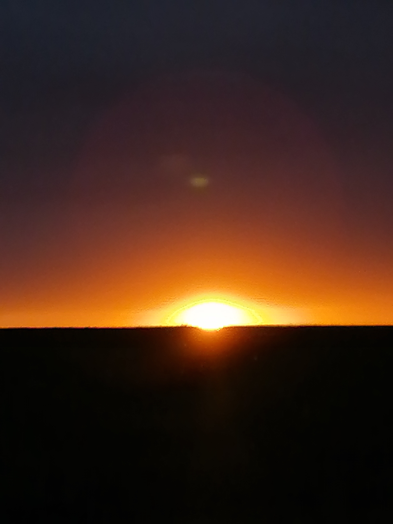 The image depicts a vibrant sunrise or sunset over a flat, dark horizon. The sky is characterized by a dramatic gradient of colors, transitioning from a deep, dark purple-blue at the top to a brilliant, intense orange and yellow directly above the horizon. The sun, a luminous white-yellow sphere, is positioned centrally, just above the stark, black horizon line, casting a strong glow that illuminates the lower atmosphere.
The horizon itself is a perfectly straight, sharp line, beneath which the entire landscape is rendered in deep silhouette, appearing as a solid black mass with no discernible features. This suggests a vast, open, and possibly flat terrain such as a plain or the sea. Subtle atmospheric haze or lens flares are visible as faint, lighter spots in the upper part of the sky, particularly one directly above the sun's position.
The image captures the serene and dramatic beauty of dawn or dusk, with the intense contrast between the radiant sky and the silhouetted foreground. There are no people, objects (other than the sun and landscape), or visible text in the frame. The scene conveys a sense of peaceful solitude and natural grandeur.