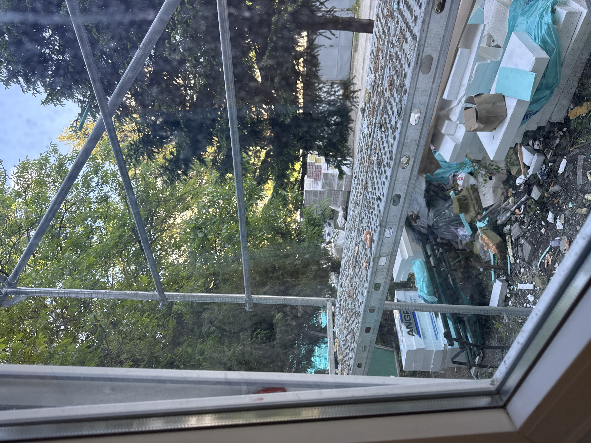 The image, possibly taken through a window, shows an outdoor scene in Lovech, Bulgaria. A metal scaffold partially obscures the view of a yard filled with construction debris. This debris includes white styrofoam blocks, fragments of insulation, trash bags, and other discarded materials. Beyond the immediate clutter, trees and greenery are visible. The image also shows the corner of a white or light-colored building or structure, possibly a house. The location is Lovech, Bulgaria.