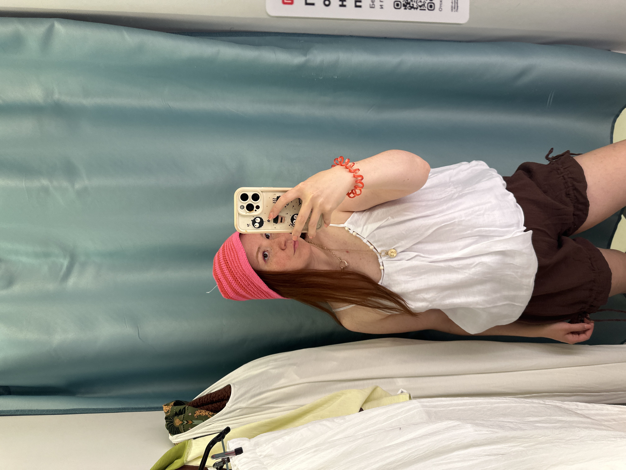 A woman is taking a selfie in what appears to be a dressing room. She is wearing a pink knitted hat, a white tank top, and brown shorts. She is holding a smartphone with a cream-colored case. She is also wearing a red bracelet and a necklace. In the background, there is a light blue curtain and other clothes hanging on a rack. Based on the information available, it is not possible to determine the location in the city of Nizhny Novgorod, Russia, or any other specific location.