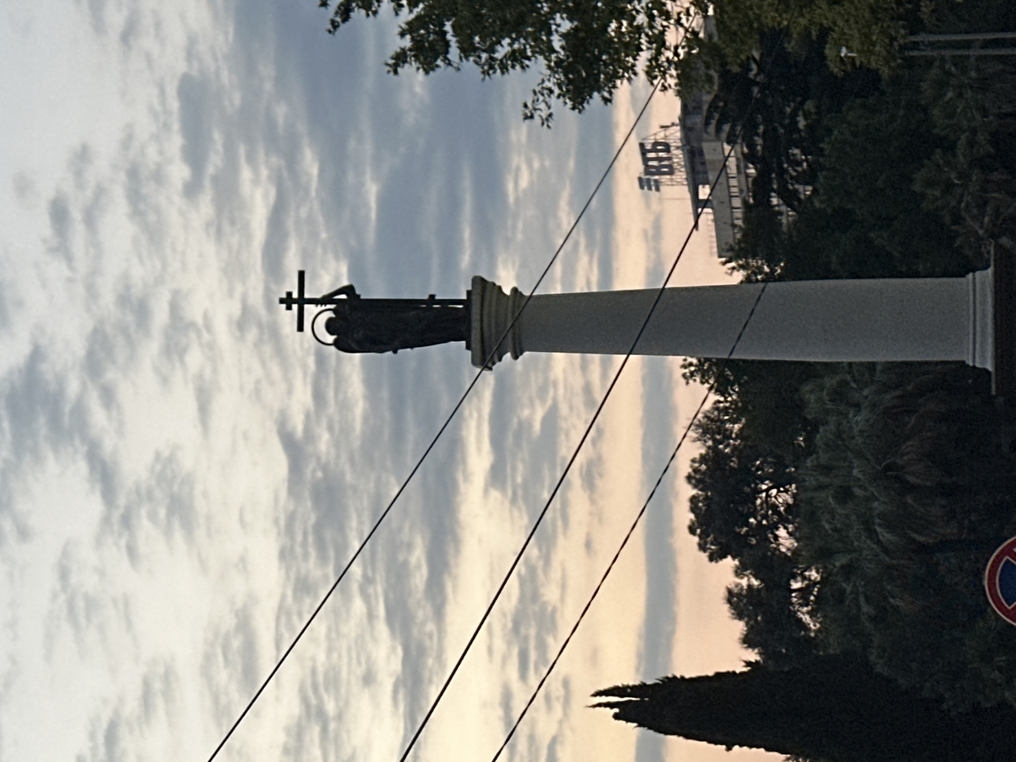 A silhouetted statue of an angel holding a cross stands atop a fluted column. The statue is dark and its features are indistinct against the overcast sky. The column is light gray and has a simple capital. In the background, a building with the letters "ВТБ" is visible. There are several trees in the foreground and midground, their foliage dark and dense. Power lines crisscross the sky above the scene. The overall lighting suggests it is either early morning or late evening.
