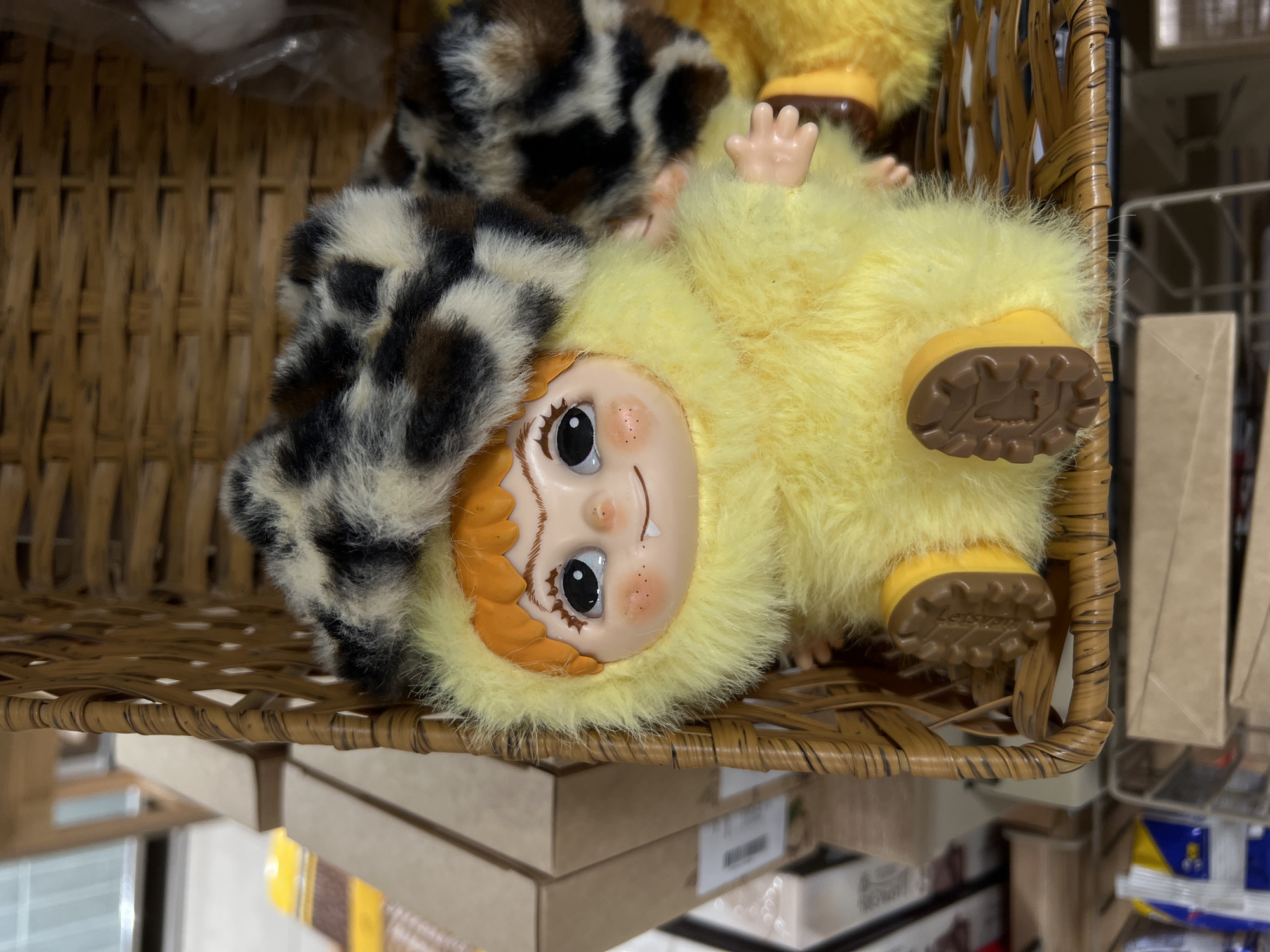 A close-up shot shows a doll in a wicker basket. The doll has a plastic face with painted features, including brown eyes, a small smiling mouth, and reddish-orange hair. It is wearing a yellow, furry costume with matching yellow boots that have brown soles. A second item, covered in black and white spotted fur, is partially visible behind the doll. The basket is sitting on a shelf, with boxes and other items visible below and to the side. The setting could be a store in Moscow, Russia, given the possibility of finding such dolls or toys in retail locations.
