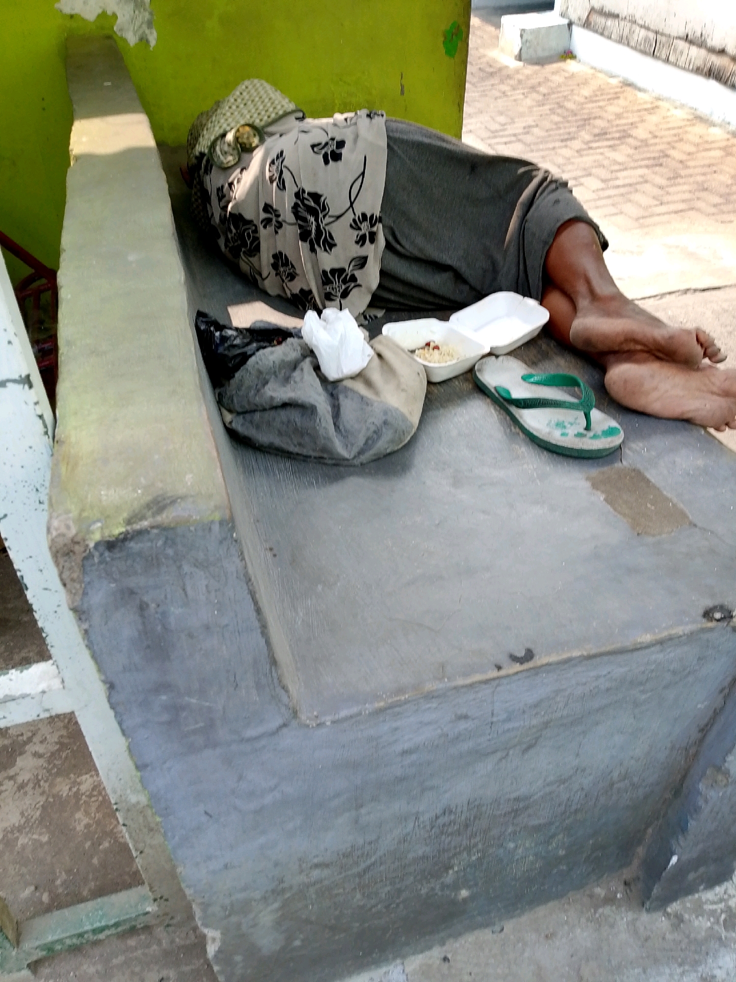 A person sleeps on a concrete bench outdoors, possibly in the city of Kediri, Indonesia. The person is covered with a patterned cloth and a dark grey blanket. Next to them is a plastic bag, a white paper container with food, and a green flip-flop. The background shows a tiled ground and a green wall.