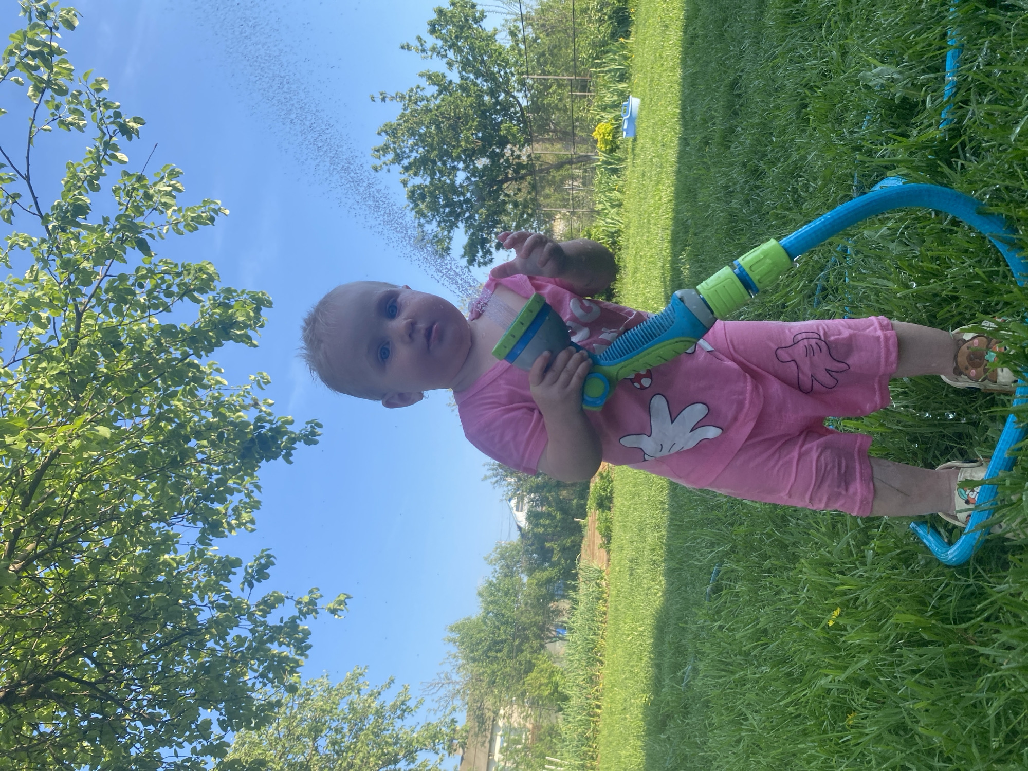 A young child, likely a toddler, is standing on a grass lawn on a sunny day. The child is wearing pink pajamas with a white cartoon hand design and sandals. The child is holding a blue and green water hose nozzle that is spraying water. Behind the child is green grass and trees under a clear blue sky. The location is Кукуи, Russia.